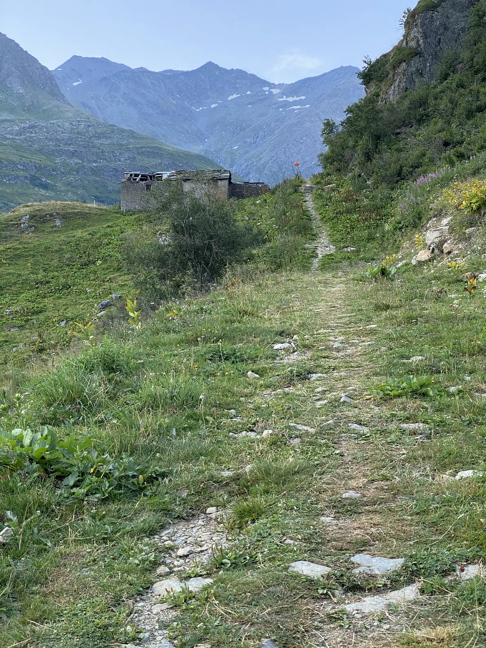 A stone-roofed ruin sits beside a stony path marked with a small red trail flag, snow-streaked mountains filling the skyline beyond.