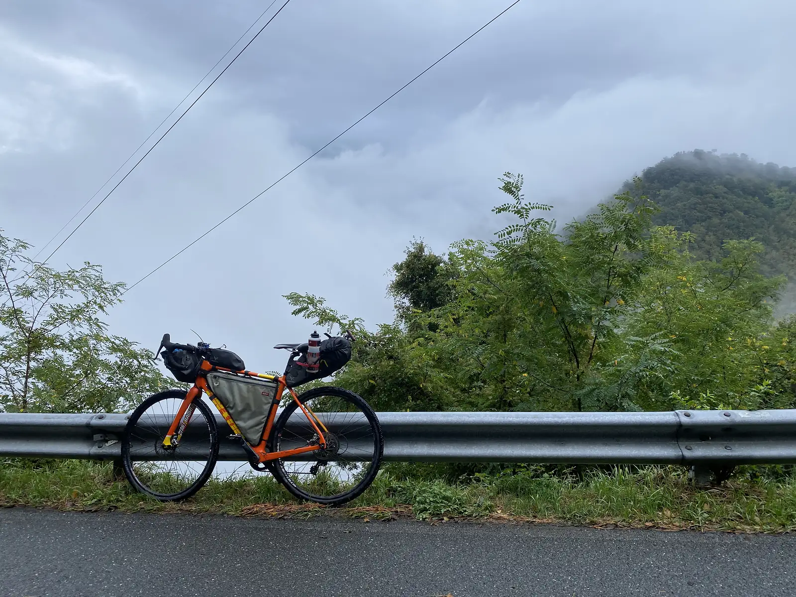 A loaded orange gravel bike rests against a steel crash barrier on a misty mountain road, forested hills rising beyond.
