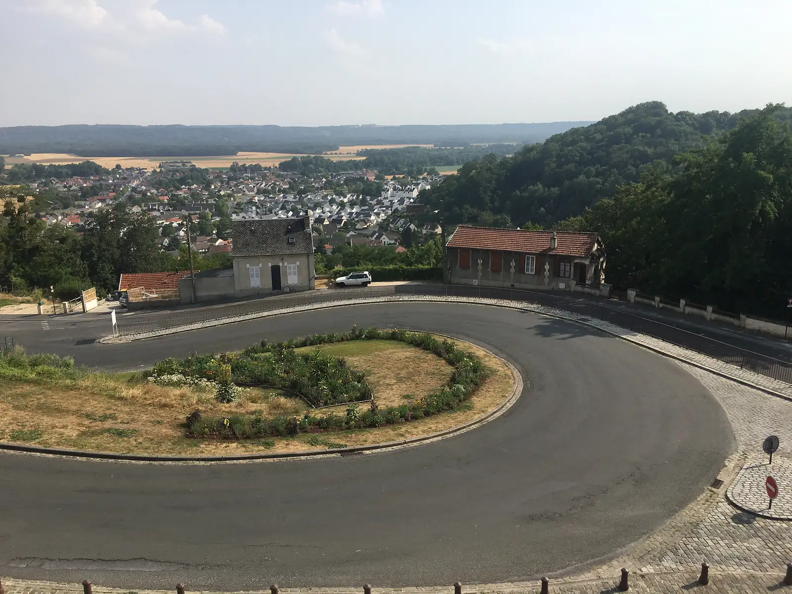 A road curves around a planted roundabout at the edge of the hilltop town, with terraced houses and wooded hills descending into the valley beyond.