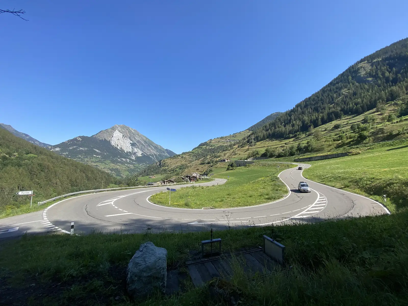 A sweeping hairpin bend on an alpine road, with a car rounding the curve, a signpost to Fornex, and mountains rising behind a scattering of chalets.