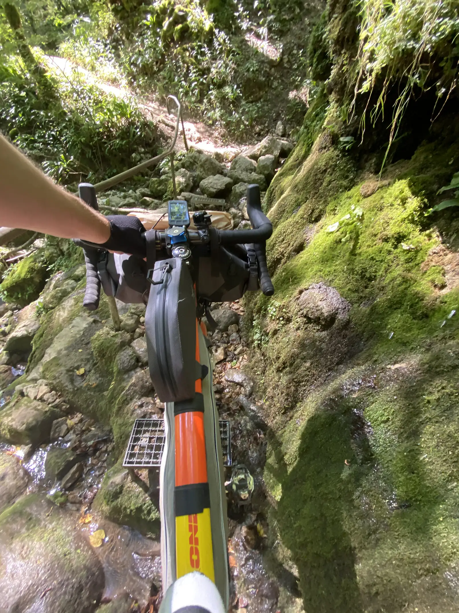 Handlebars laden with bikepacking bags point down a boulder-strewn streambed crossed by metal grating, mossy rock walls rising on either side.
