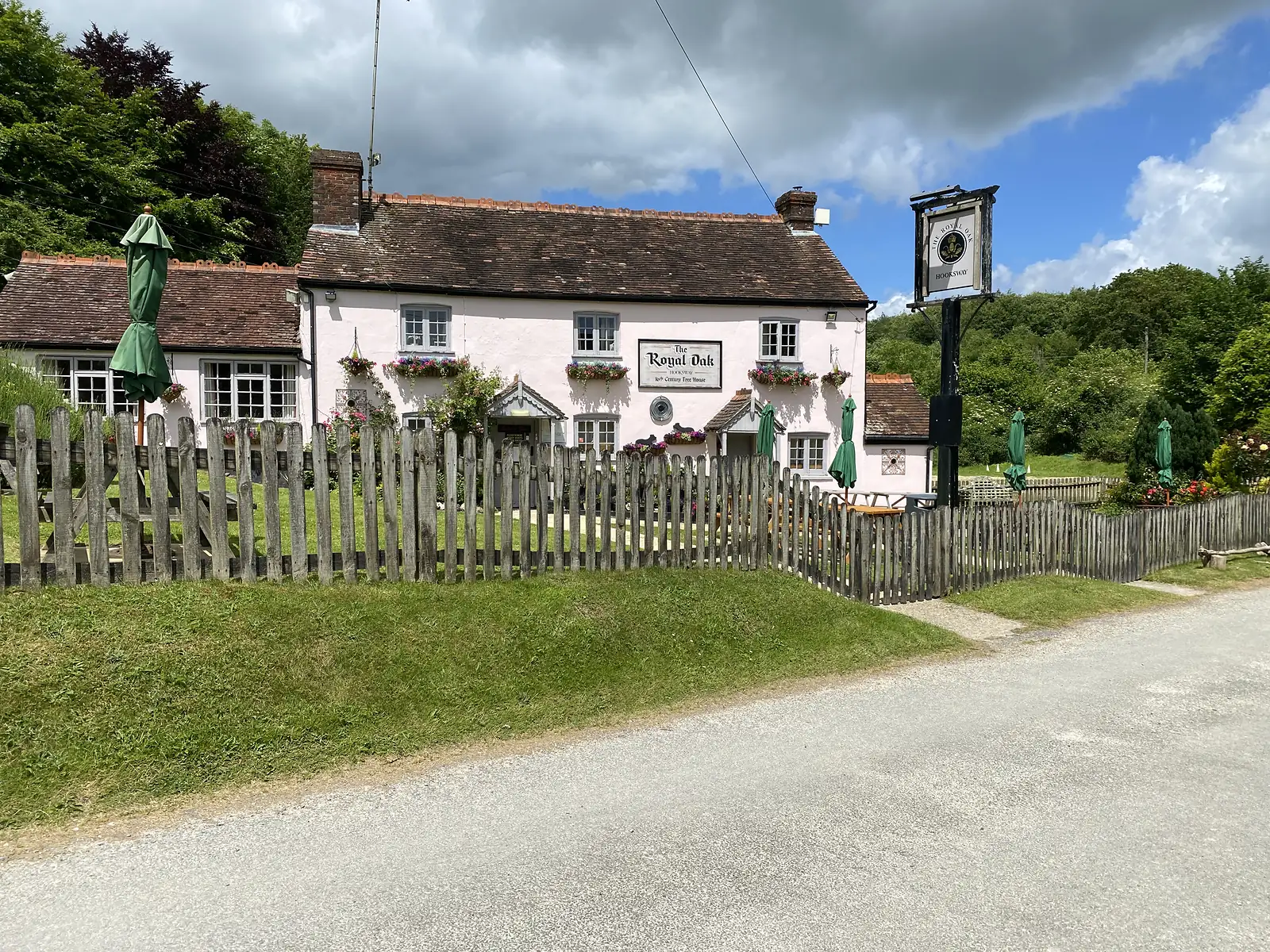 A pink-washed tile-roofed country pub lettered "The Royal Oak" behind a picket fence, with hanging baskets and furled green parasols.