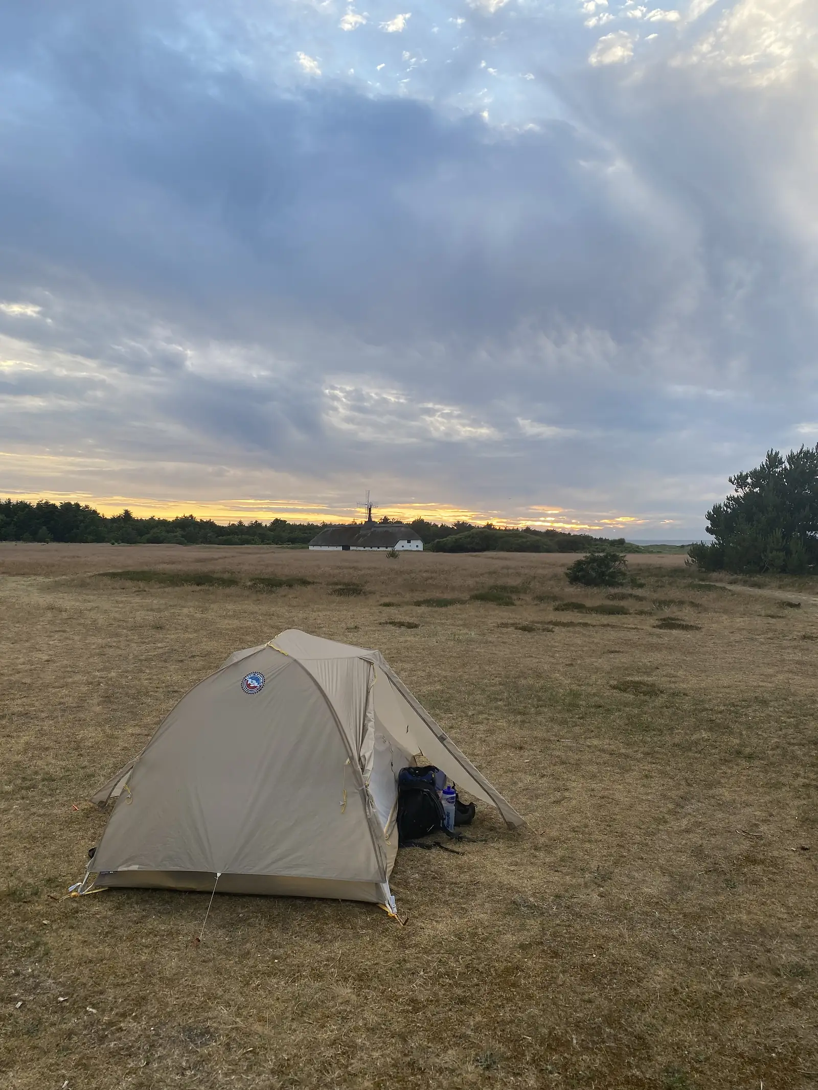 A small tent pitched on dry heath at dusk, with a thatched windmill farmstead silhouetted on the horizon against a yellow sunset.