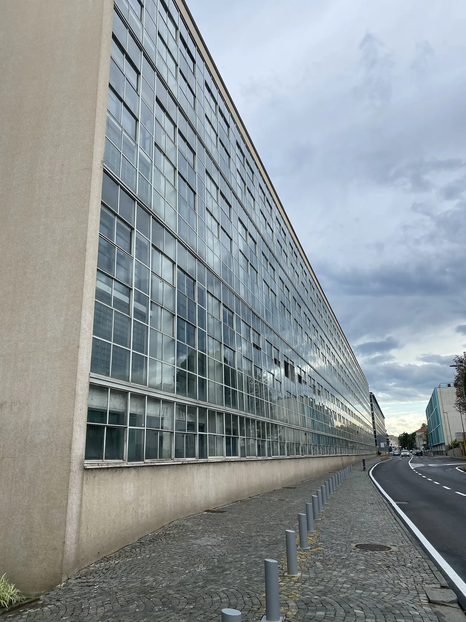 The long glazed curtain-wall facade of a former factory building stretching into the distance alongside a road, its many small window panes reflecting a cloudy sky.
