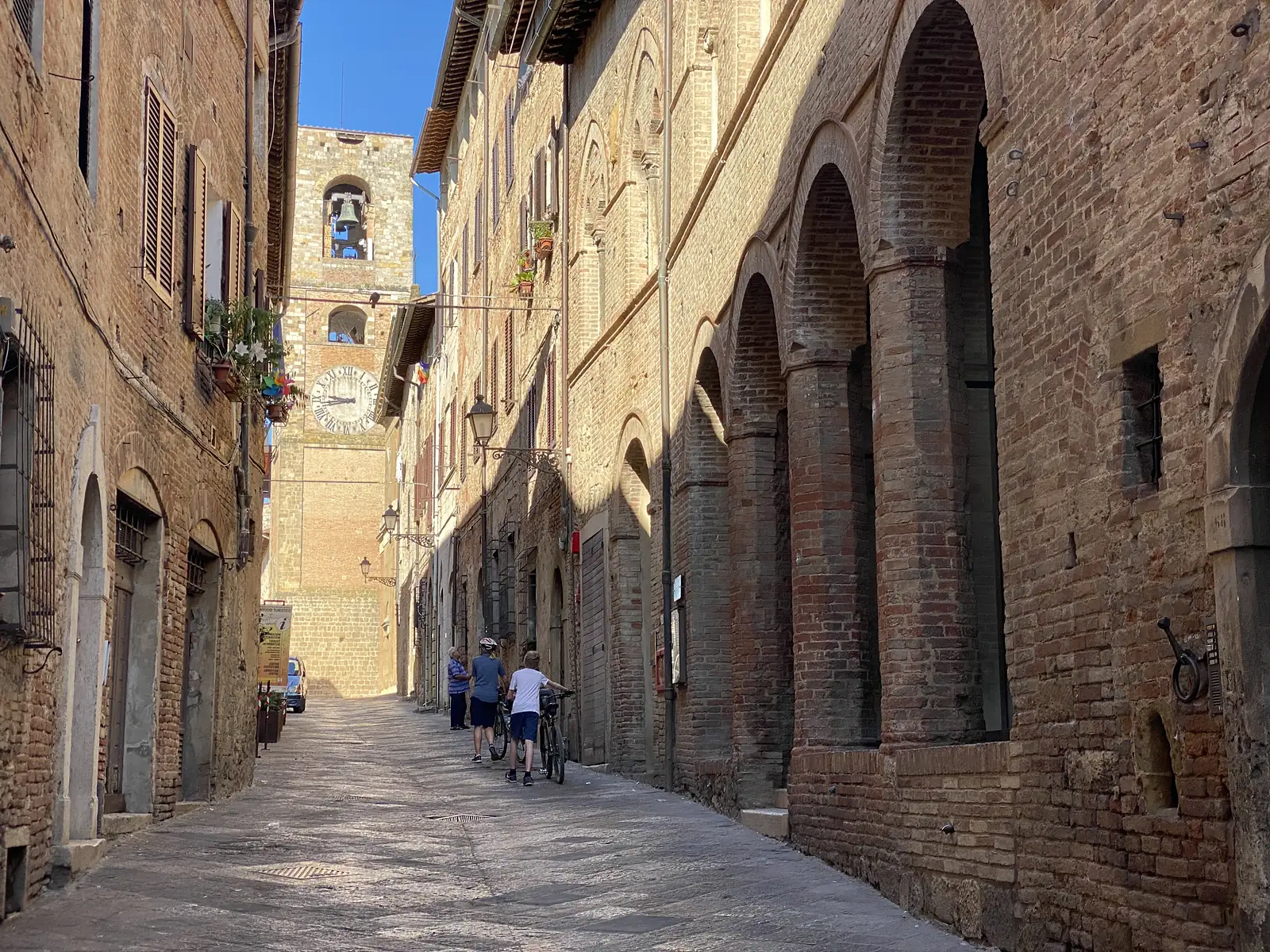 Cyclists push their bikes down a narrow brick-paved lane lined with an arcaded portico, heading toward a clock tower with a bell.