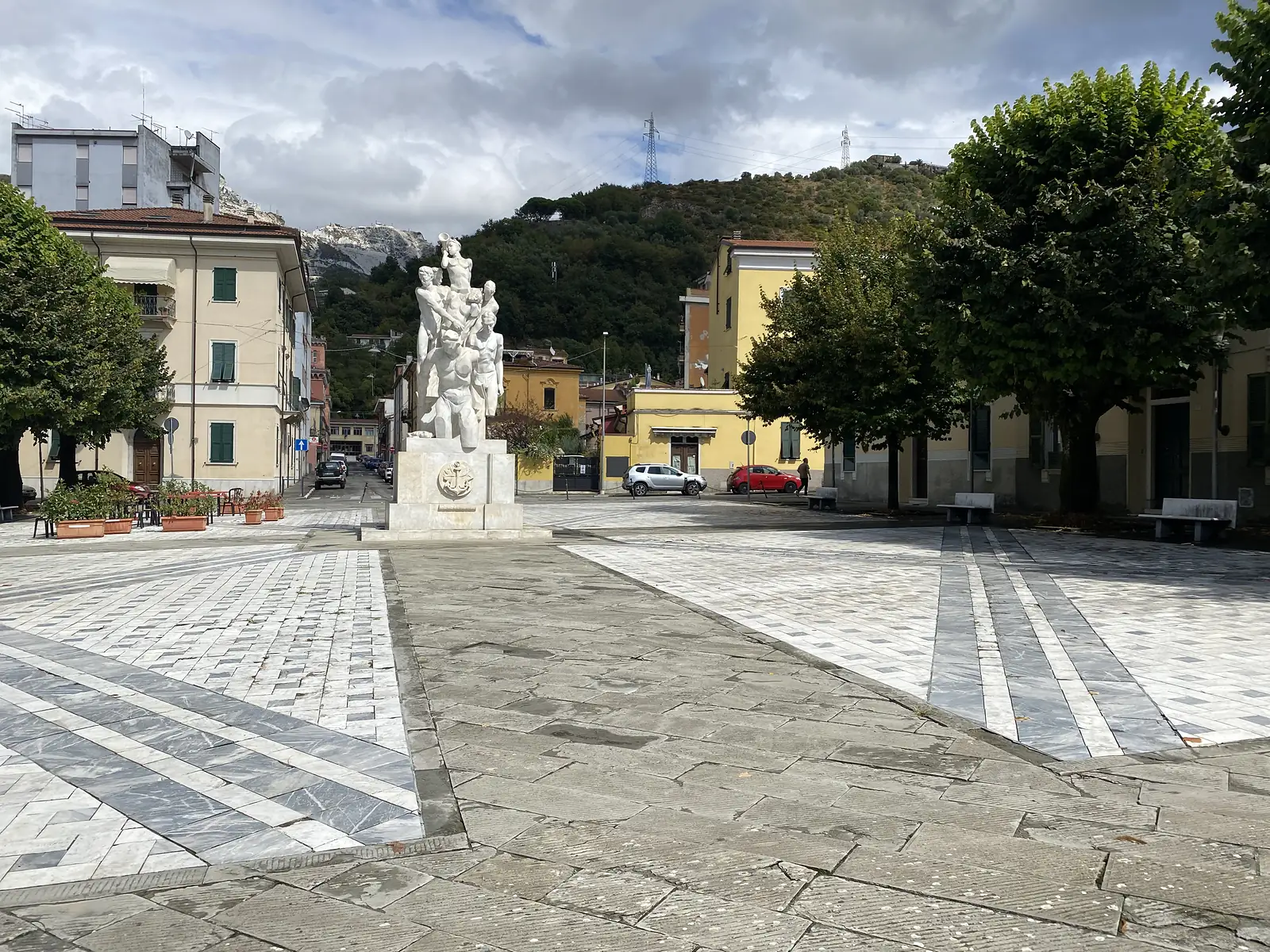 A white marble memorial of intertwined sailor figures topped by a trumpeter, its plinth bearing an anchor emblem, stands in a marble-paved piazza with quarried mountains visible behind the surrounding buildings.