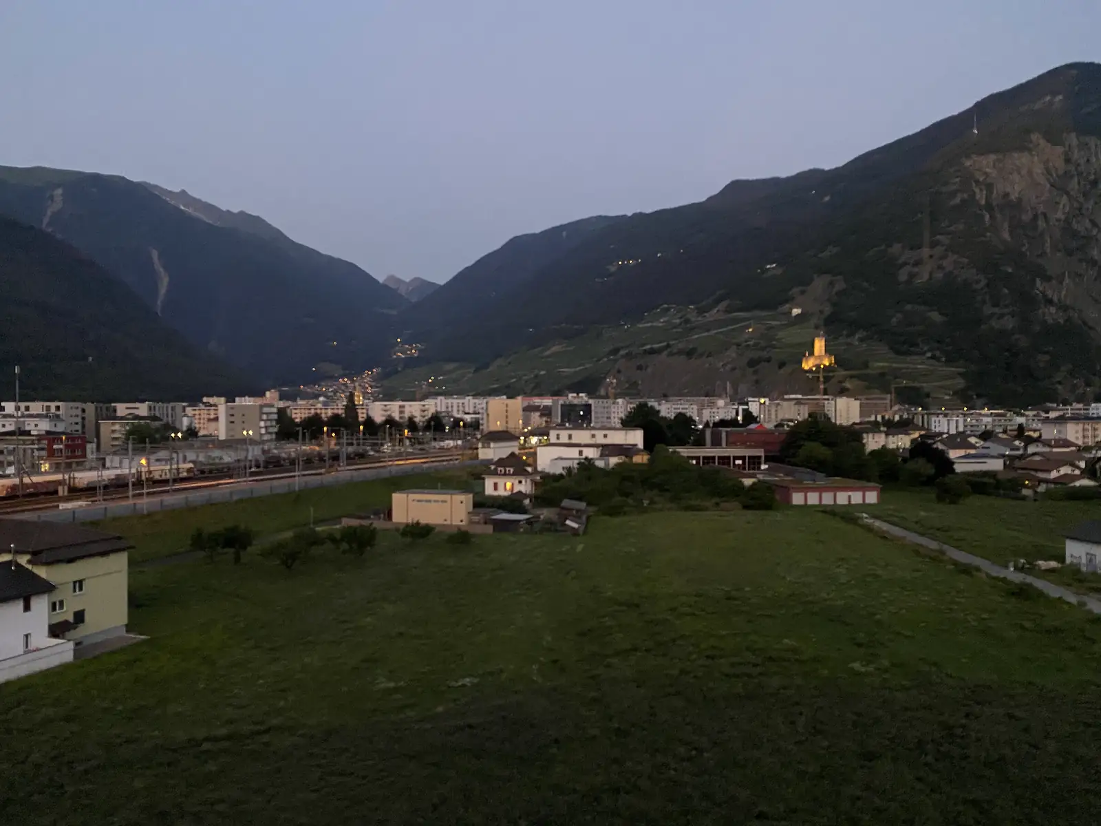 A Swiss valley town lit up at dusk, seen from a grassy hillside above a railway line, with a floodlit hilltop tower and dark mountains rising behind.