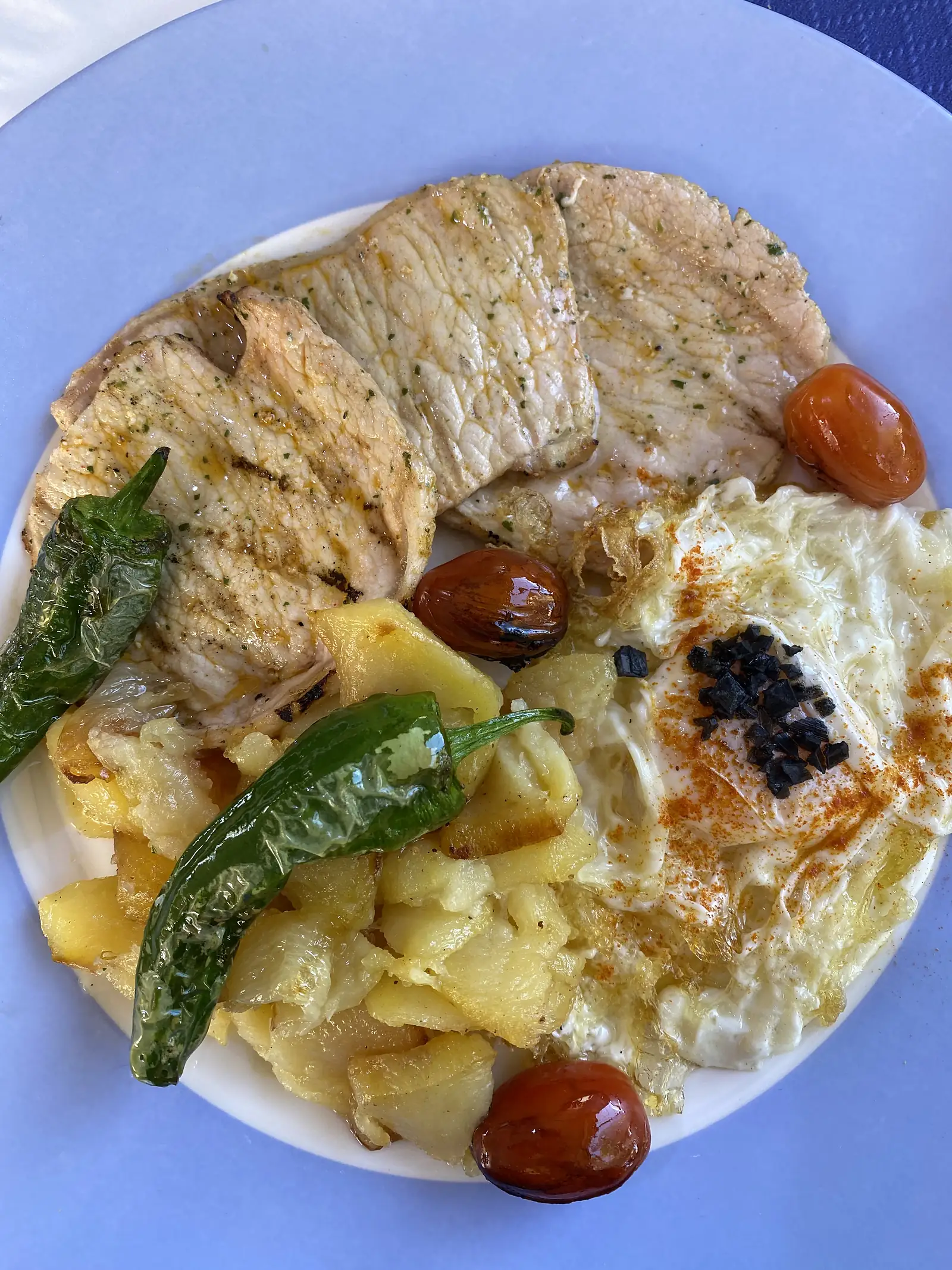 Grilled pork loin steaks with fried potatoes, a paprika-dusted fried egg, green peppers and cherry tomatoes on a plate on a blue table.