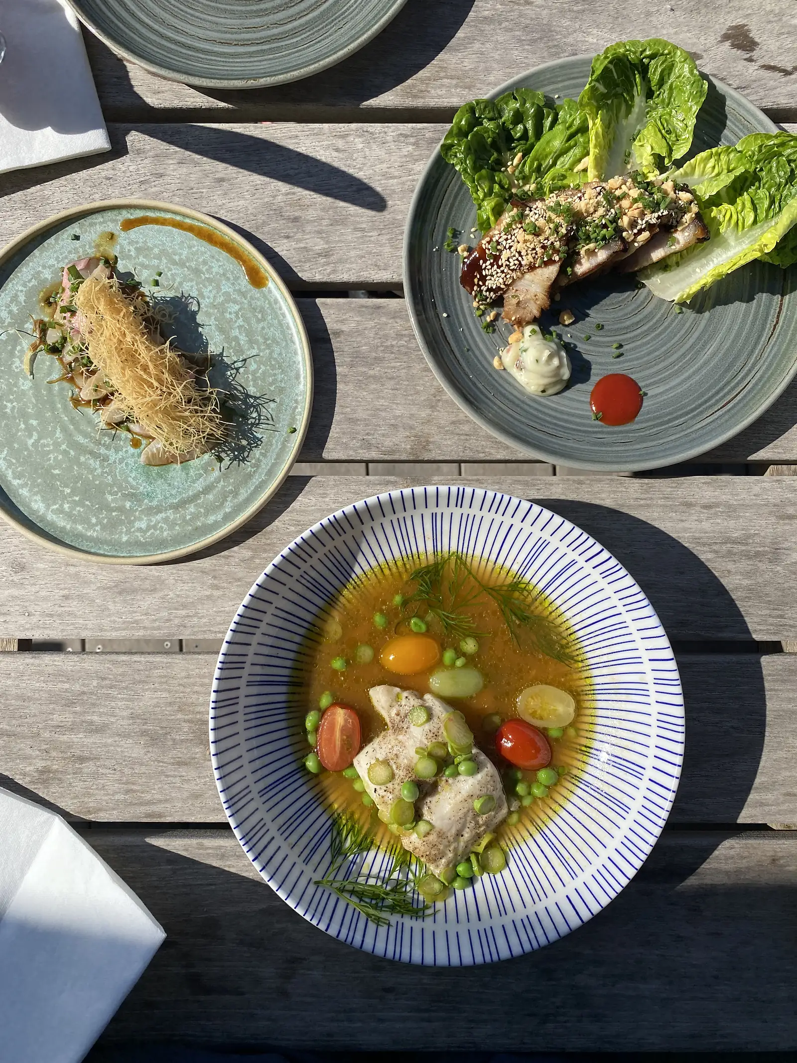 Three restaurant dishes seen from above on a weathered wooden table: fish in broth with peas and dill, sliced pork with peanuts and lettuce, and raw fish under crisp shredded pastry.