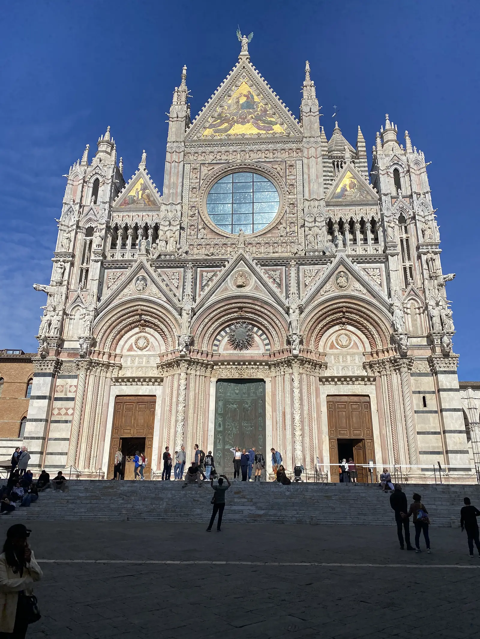 The elaborate striped marble facade of a Gothic cathedral, richly decorated with statues, spires and gold mosaic gables, with visitors gathered on the piazza below its bronze doors.