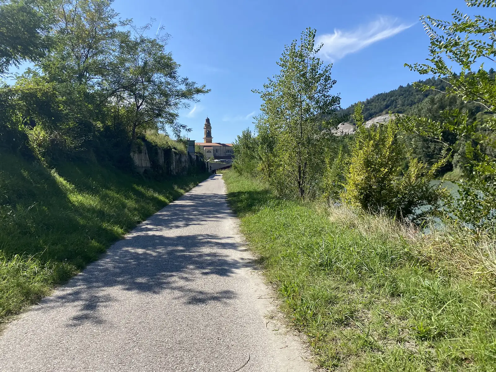A cycle path between grassy banks heads towards a village dominated by a tall church tower, with a river running alongside.