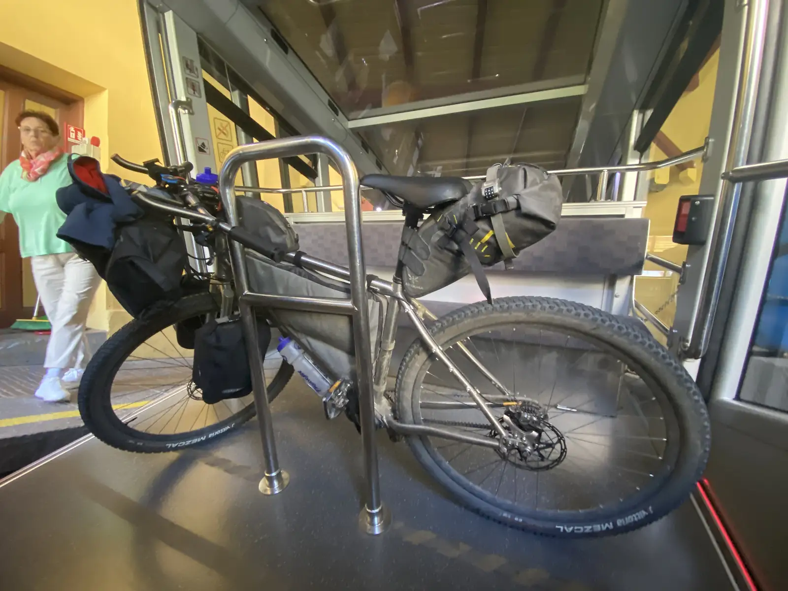 A loaded bikepacking bike stands strapped into the bike bay of a funicular car, with a passenger walking past on the platform outside.