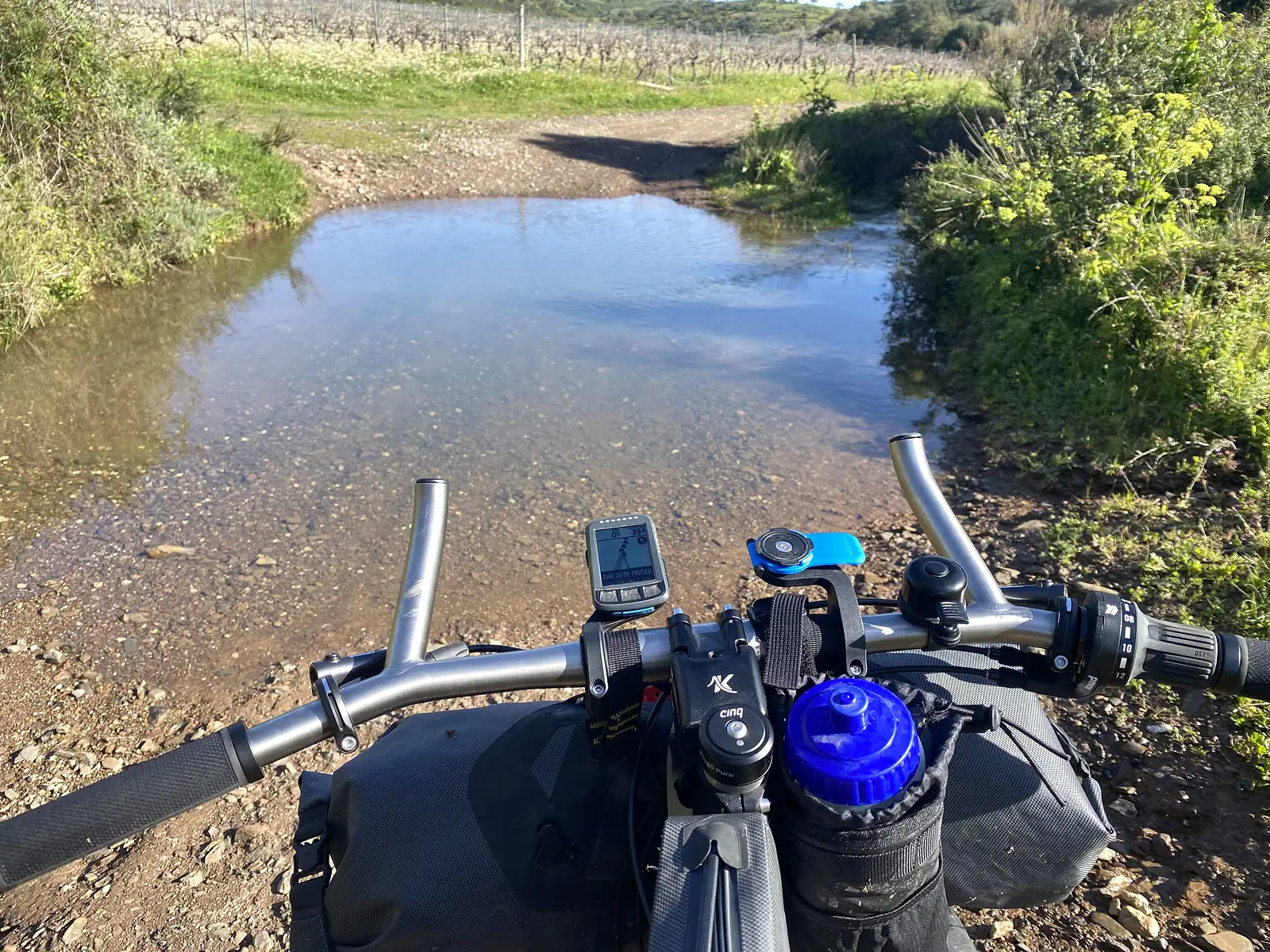 Rider's view over loaded handlebars, bar bag and computer at a wide ford where the track disappears into shallow brown water, vines climbing the slope beyond.