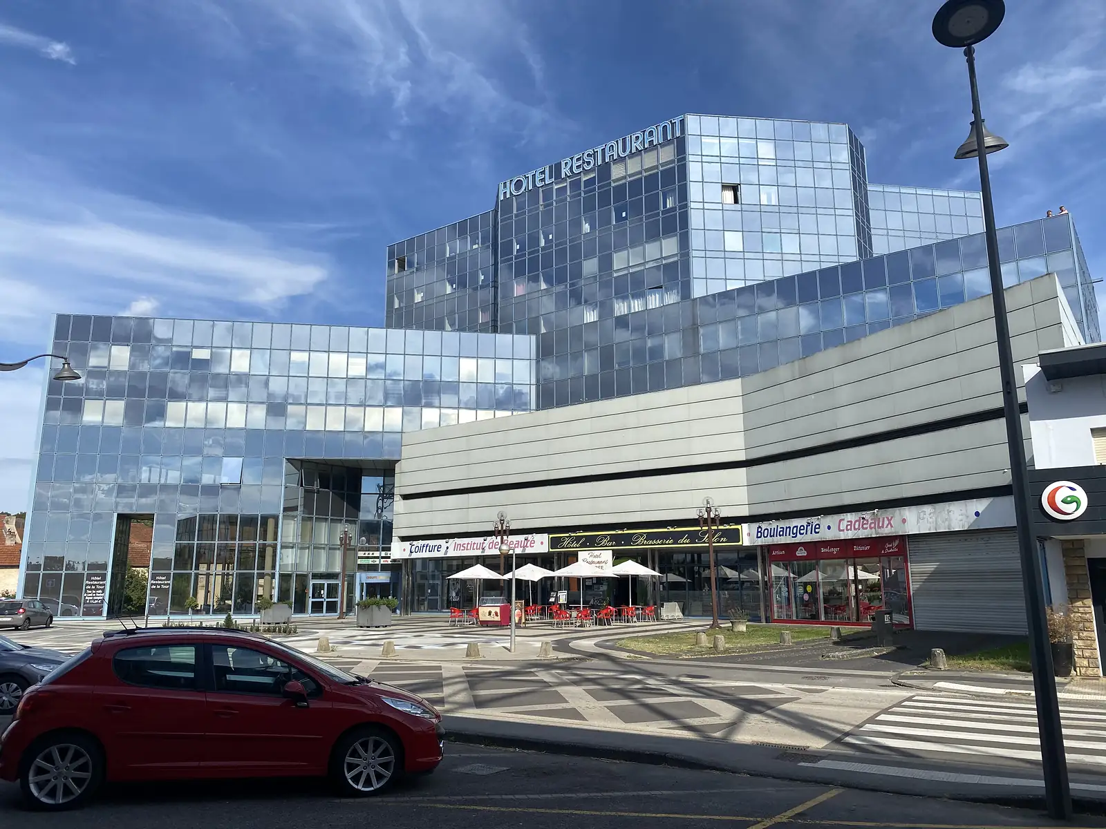 A modern glass-fronted building bearing a large 'HOTEL RESTAURANT' sign rises above a row of shopfronts including a boulangerie and hairdresser, with a red car parked in the square outside.