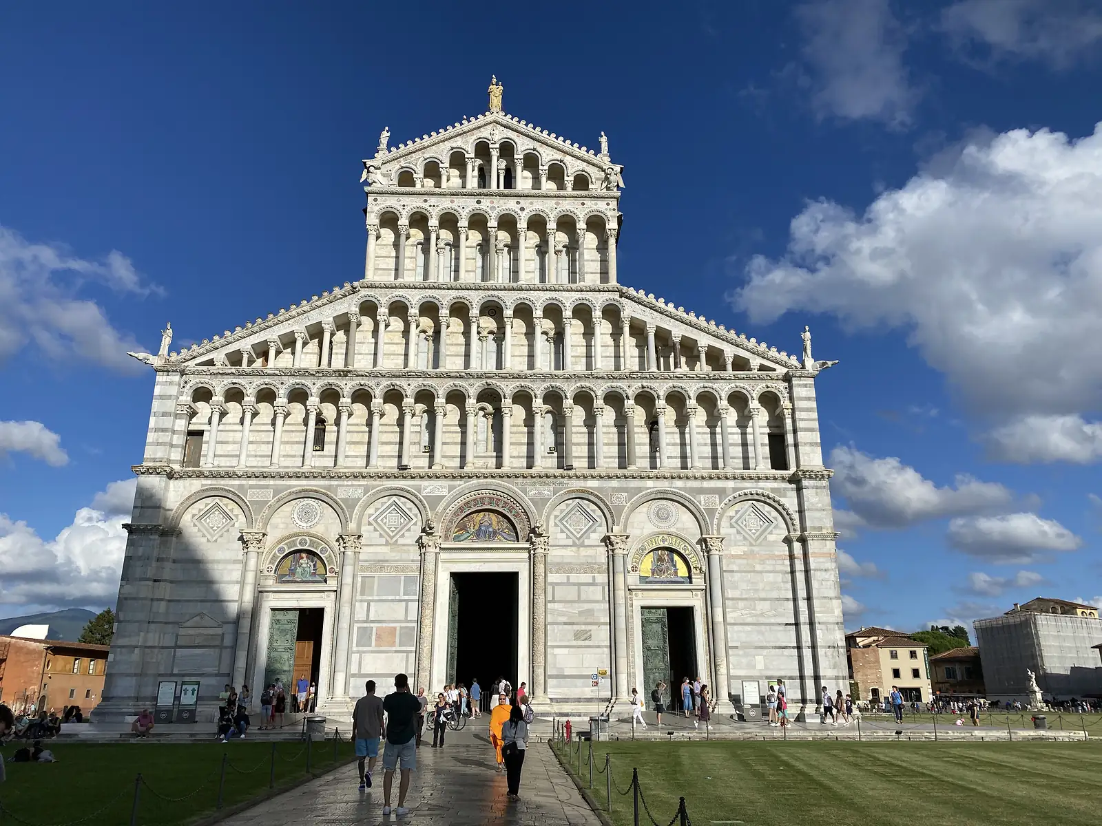 The striped marble facade of Pisa Cathedral, its tiers of blind arcading and columned loggias rising to a gilded statue at the roofline, with visitors gathered at its open doors.