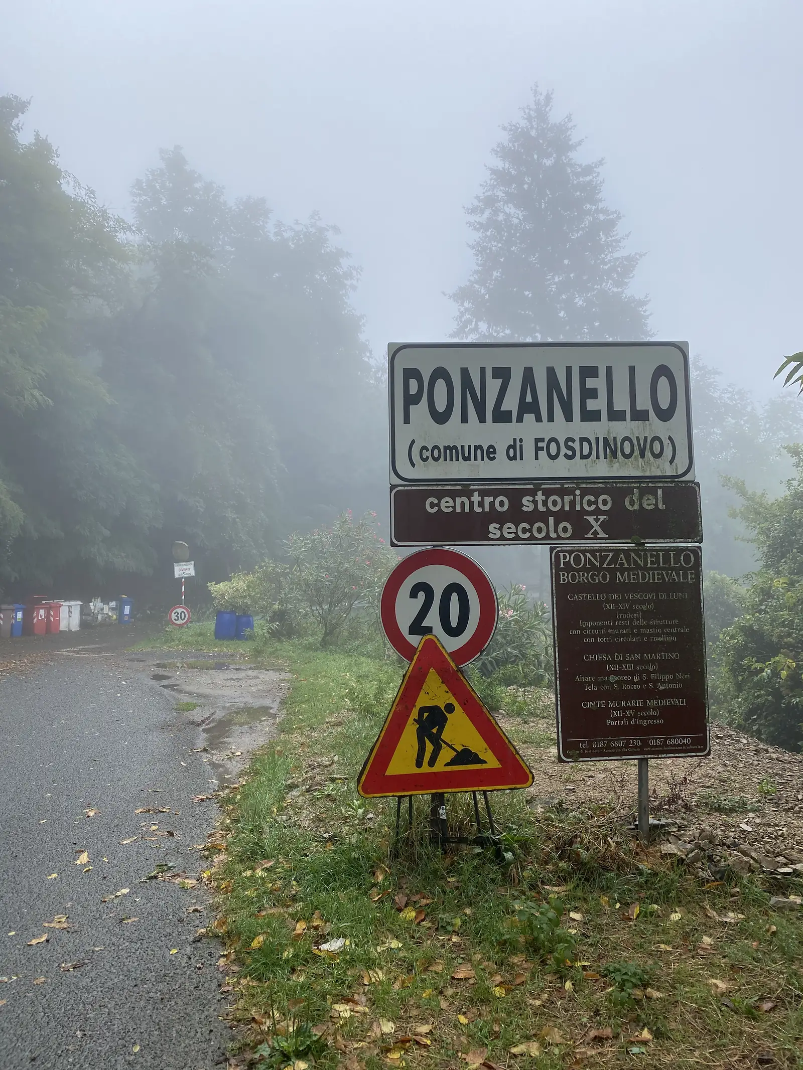 Roadside signs announce the village of Ponzanello, in the comune of Fosdinovo, noting its medieval old town dating from the tenth century, alongside a 20 km/h limit sign and a pedestrian warning triangle in thick fog.