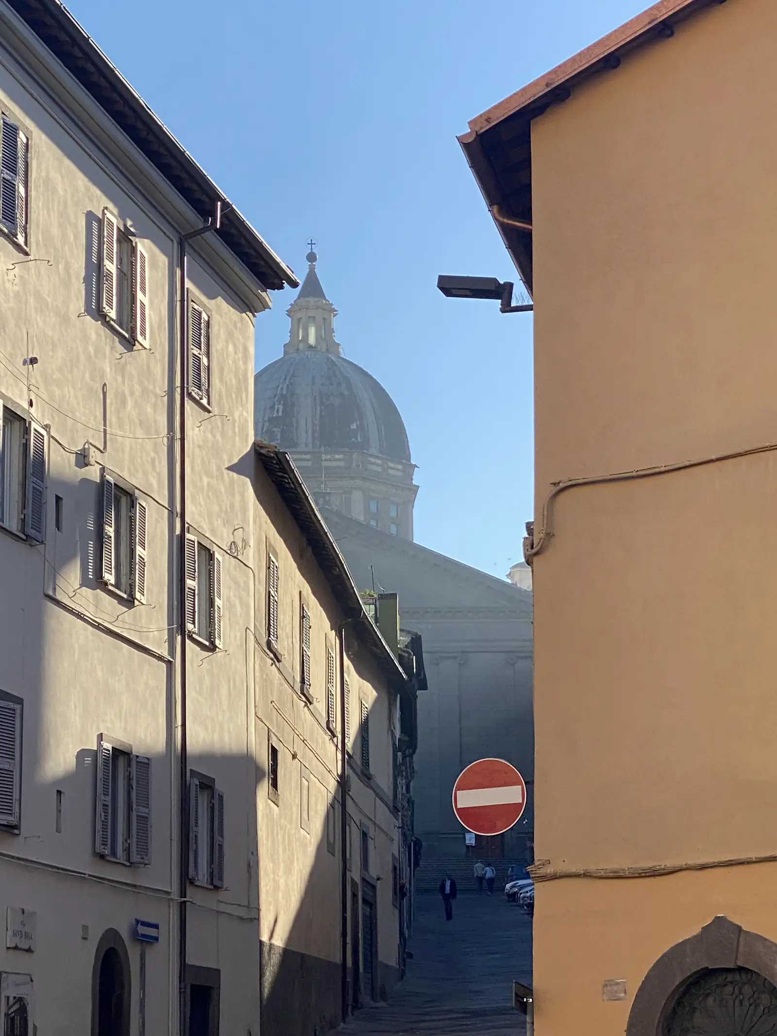 A domed church tower rising above the shuttered facades of a narrow town street, a no-entry road sign fixed to a building on the corner.