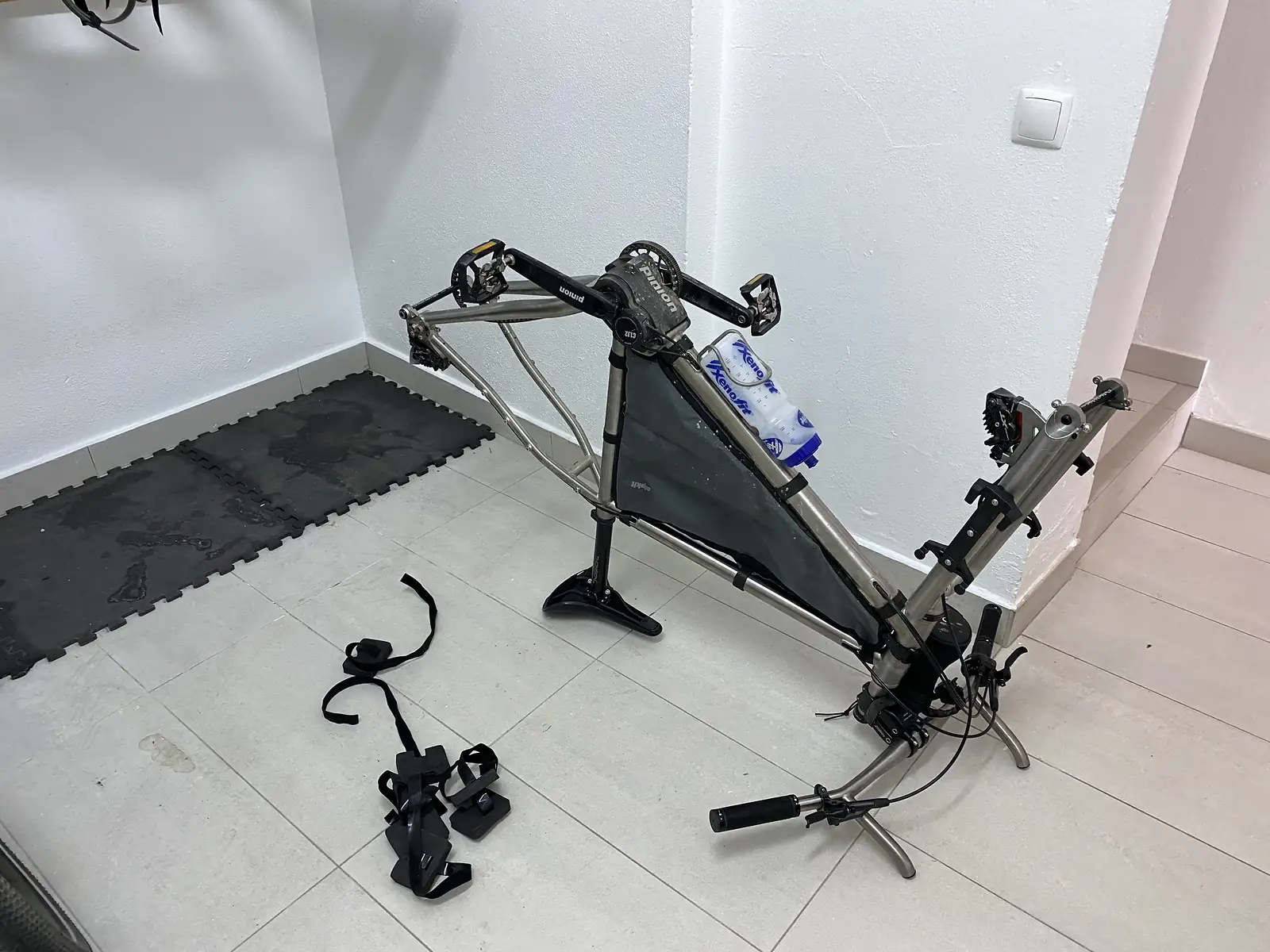 Partly stripped touring bike frame propped on a stand against a white wall on a tiled floor, frame bag still fitted, forks and bars hanging loose and pedals, straps and cargo cages laid out alongside.