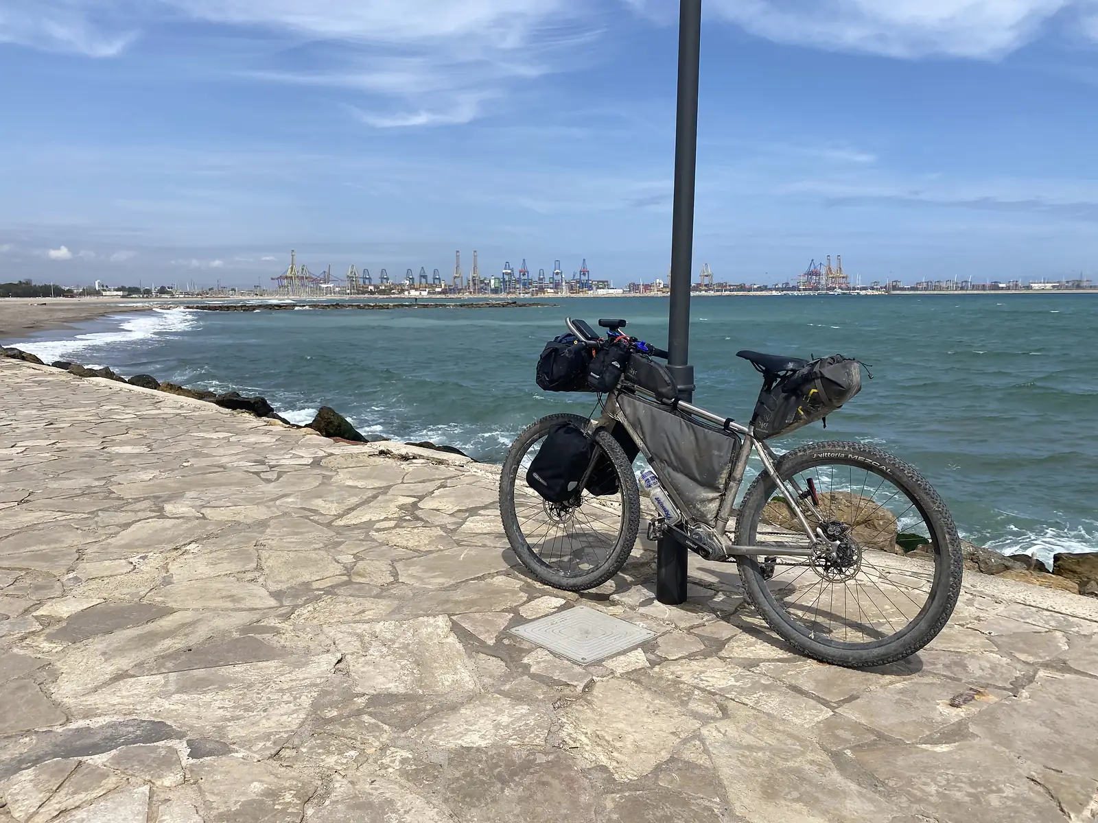 A travel-worn loaded bikepacking bike leaning on a lamp post on a stone-flagged sea wall, with a container port's cranes across the bay.
