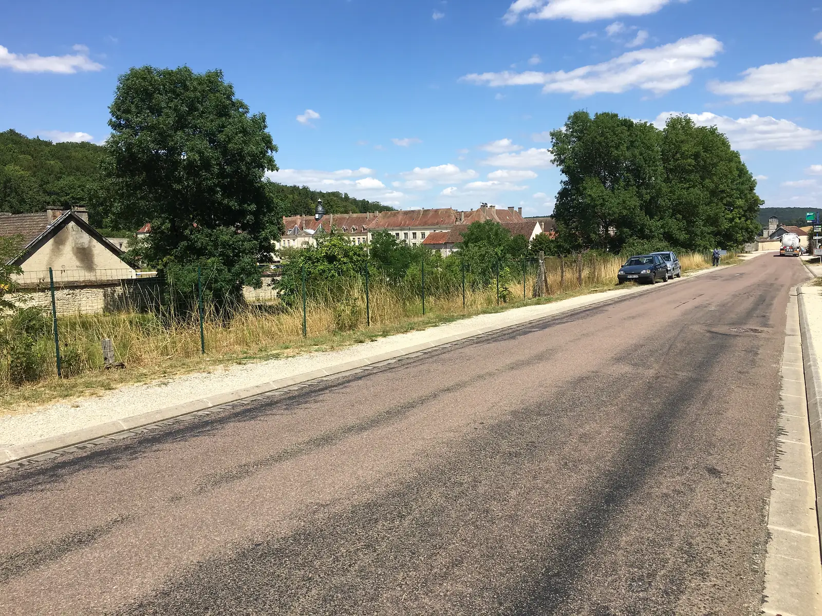 A quiet village road lined with mature trees leads towards a cluster of stone houses, with a parked car and a pedestrian in the distance.