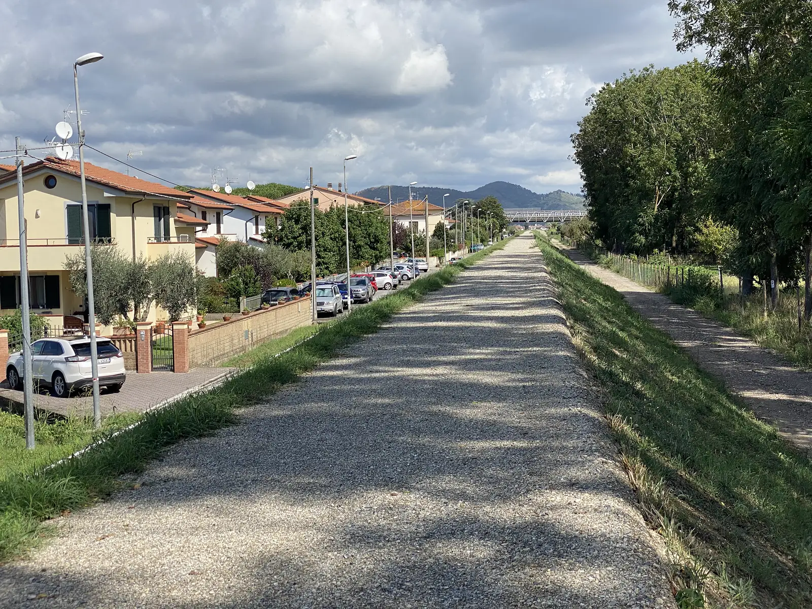 A gravel lane running past terracotta-roofed Tuscan houses towards distant hills and a railway viaduct.