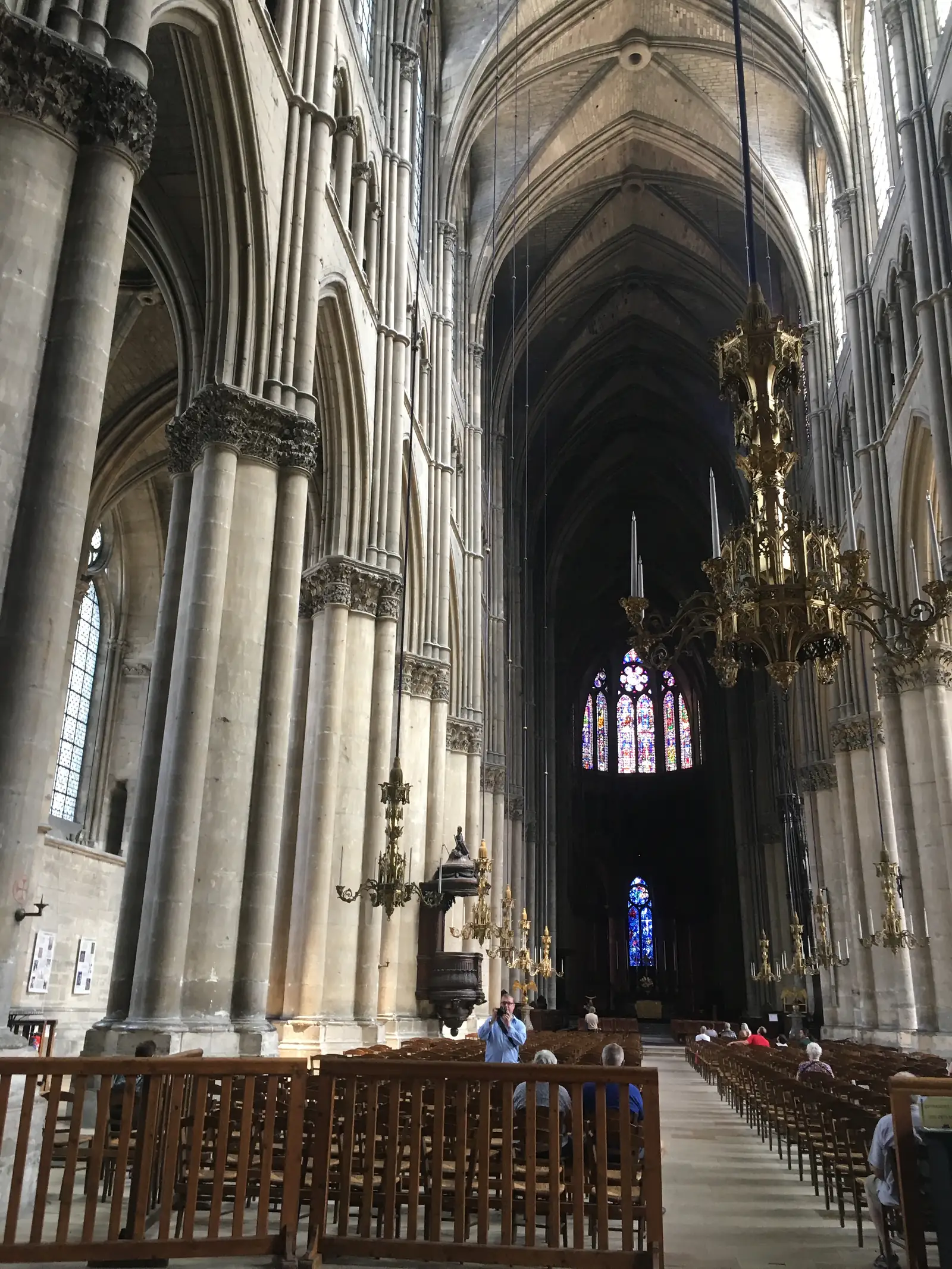 Looking up the nave of Reims Cathedral towards a glowing stained-glass rose window above the choir, gilded candelabra hanging from soaring gothic arches.