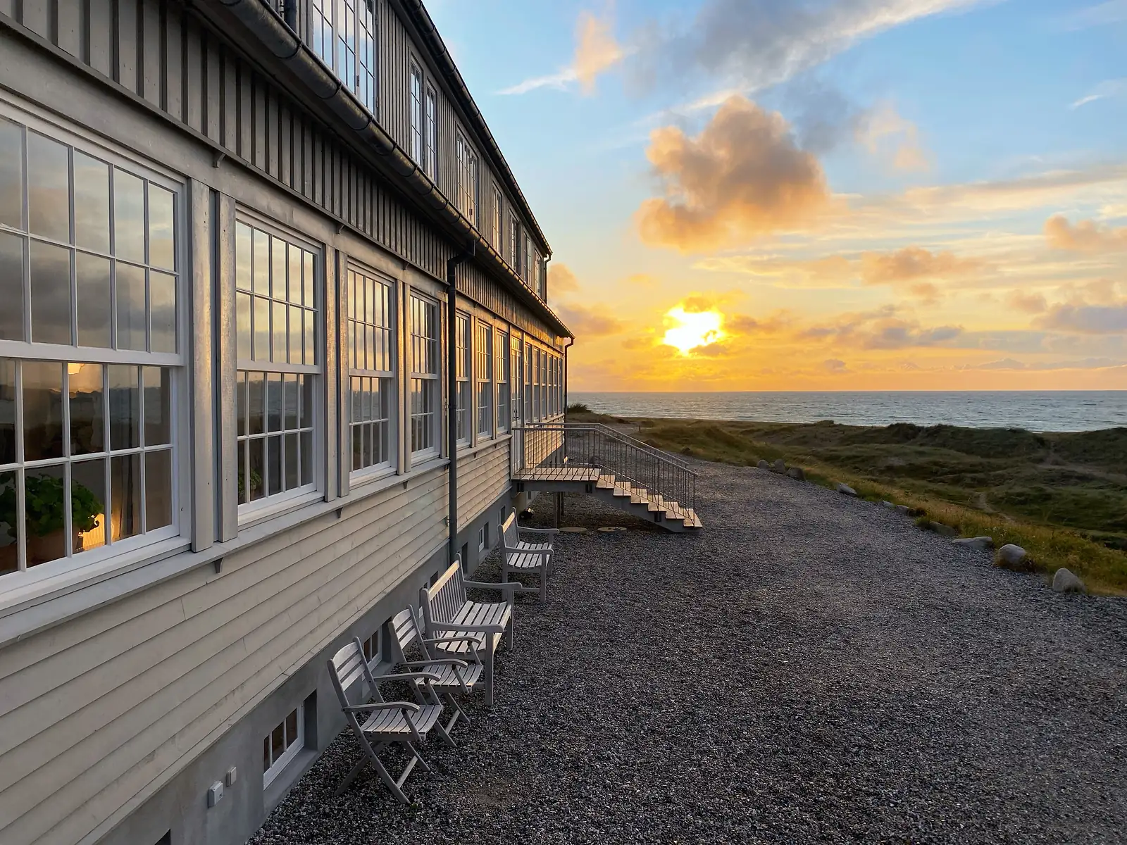 A long timber-clad building with tall many-paned windows and weathered wooden chairs on a gravel terrace above the dunes, the sun setting over the sea.