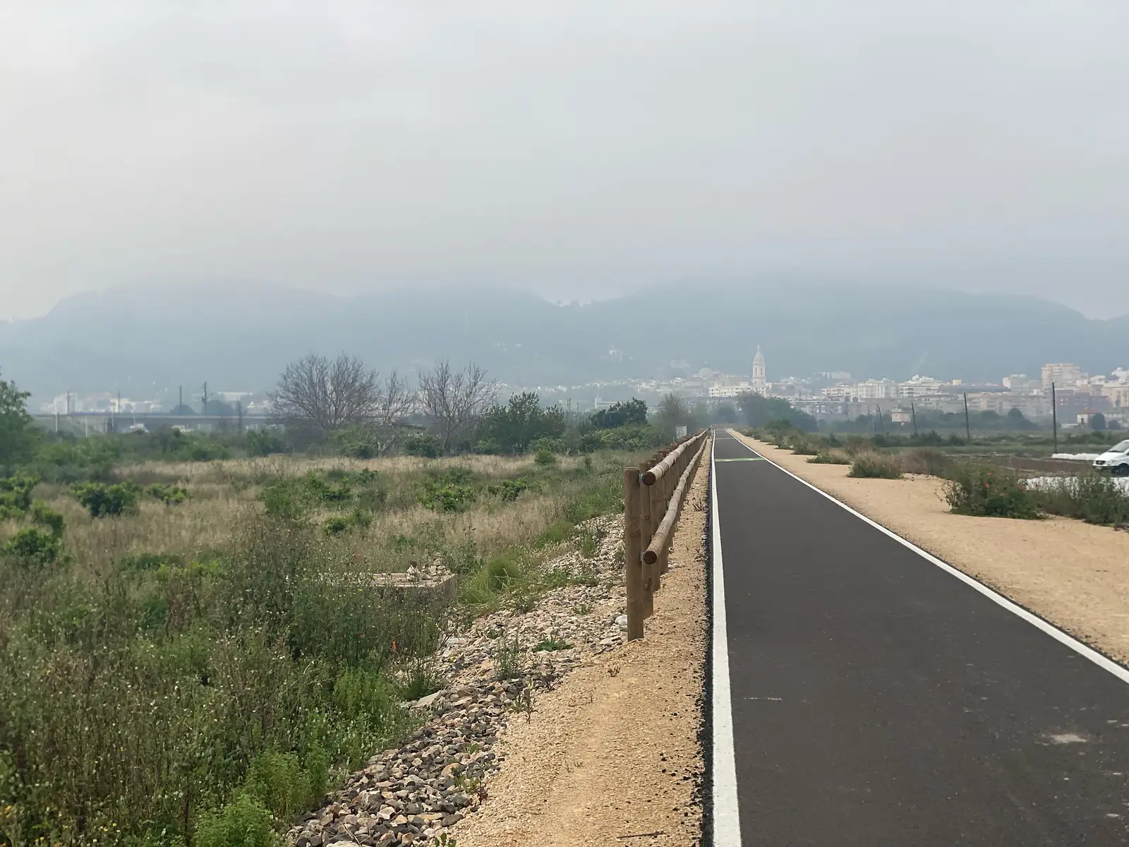 A new asphalt greenway running dead straight between orange groves and a post-and-rail fence towards a hazy city with a tall bell tower.