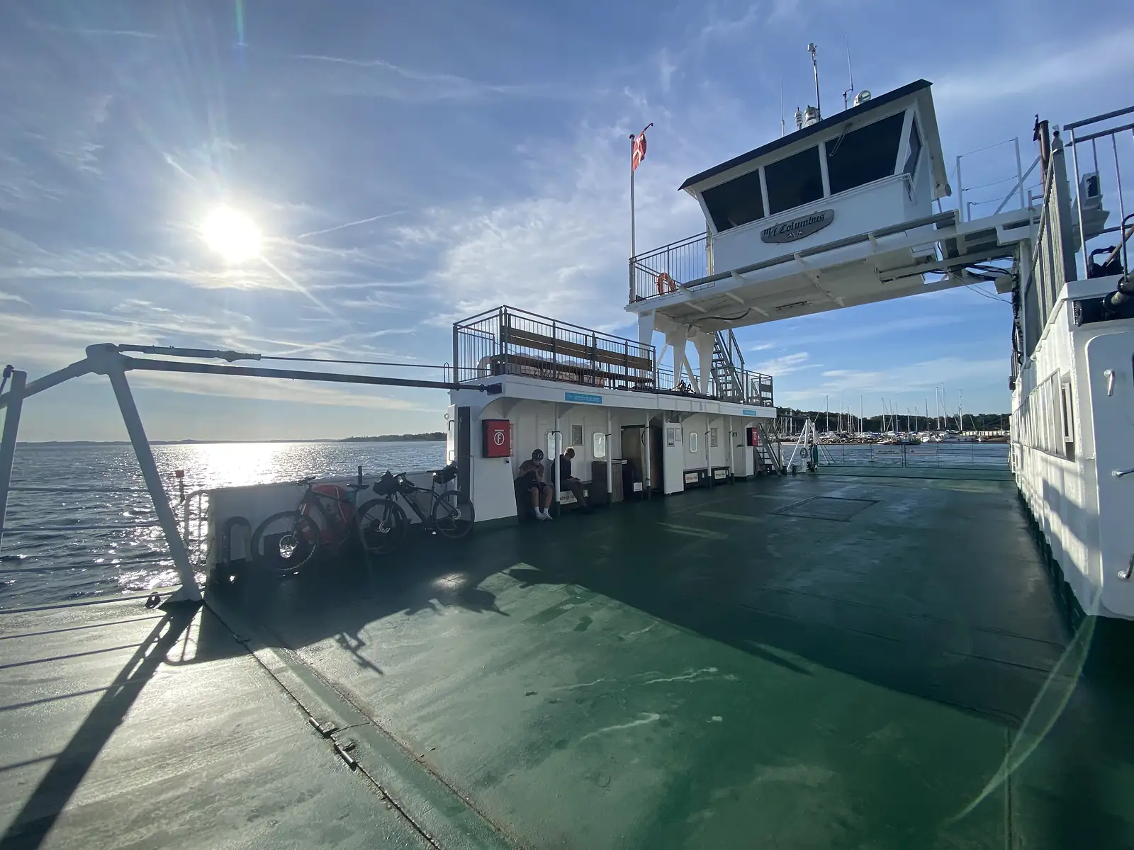 Bikes lined along the rail of a ferry's green deck in low morning sun, wheelhouse and Danish flag above.