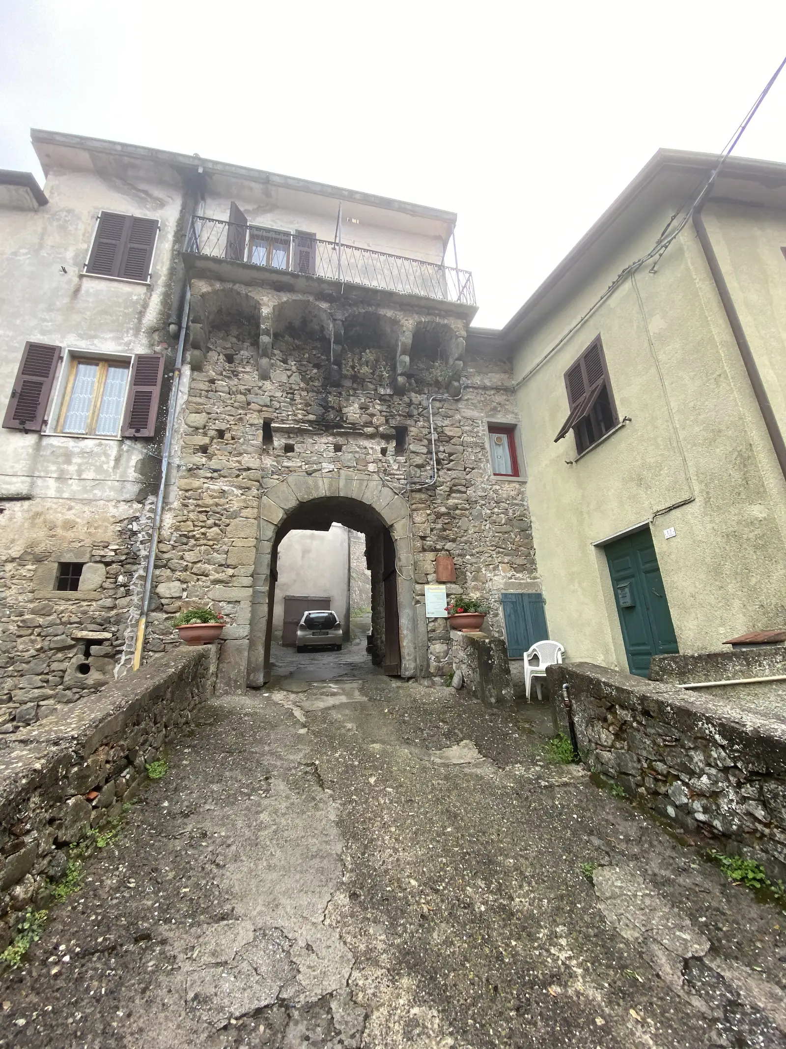 A vaulted stone archway leads from a paved lane into a small courtyard with potted plants, shuttered windows and a red door.