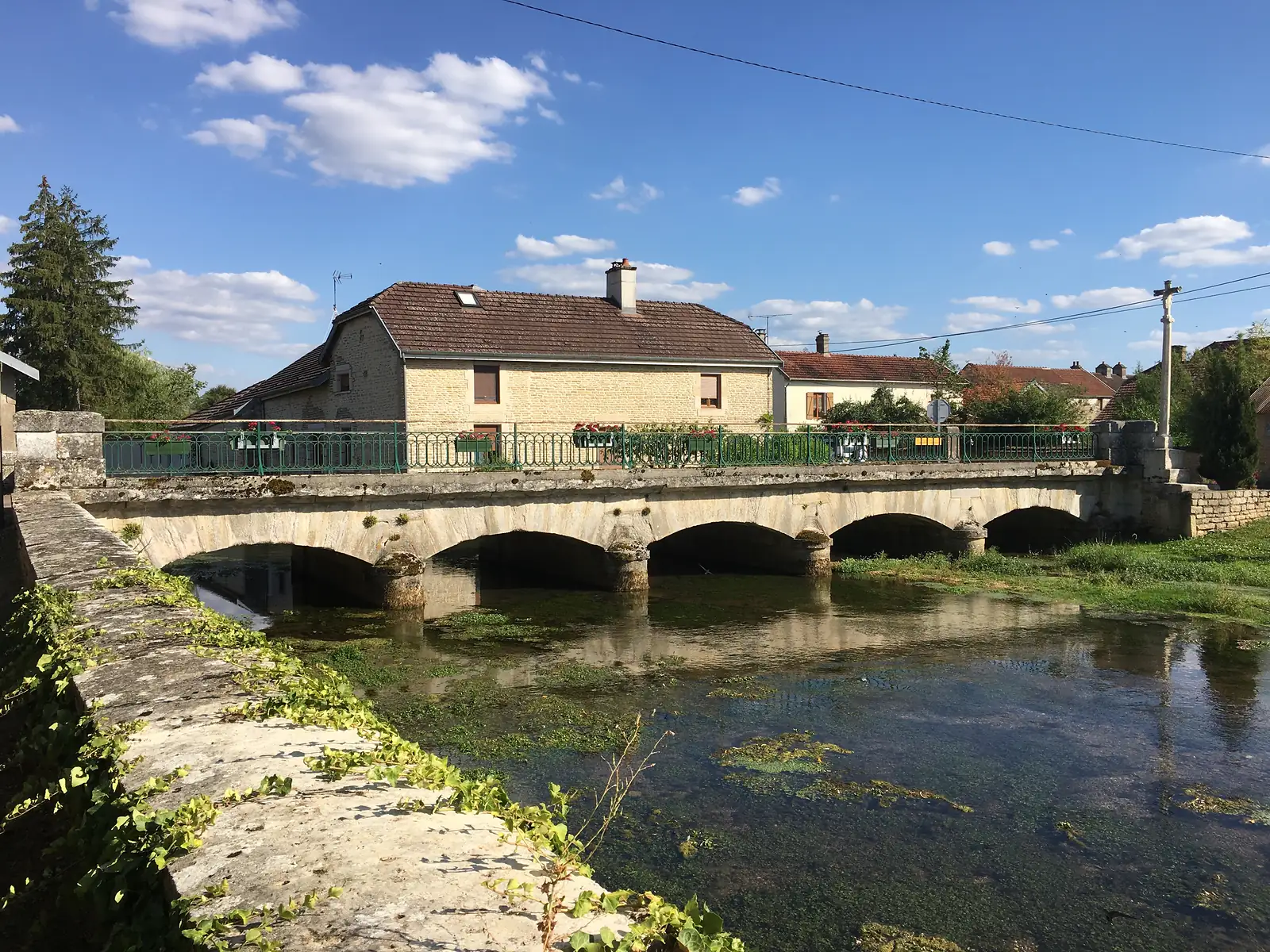 A stone-arched bridge crosses a river overhung with willows, with a wayside cross and a 'STOP' sign standing at its near end.