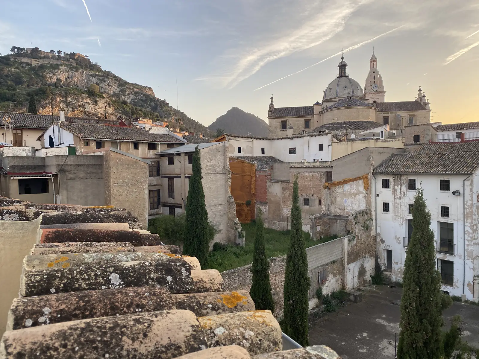 Tiled rooftops and derelict houses below a hillside castle wall, with a domed church and clock tower catching the dawn light.