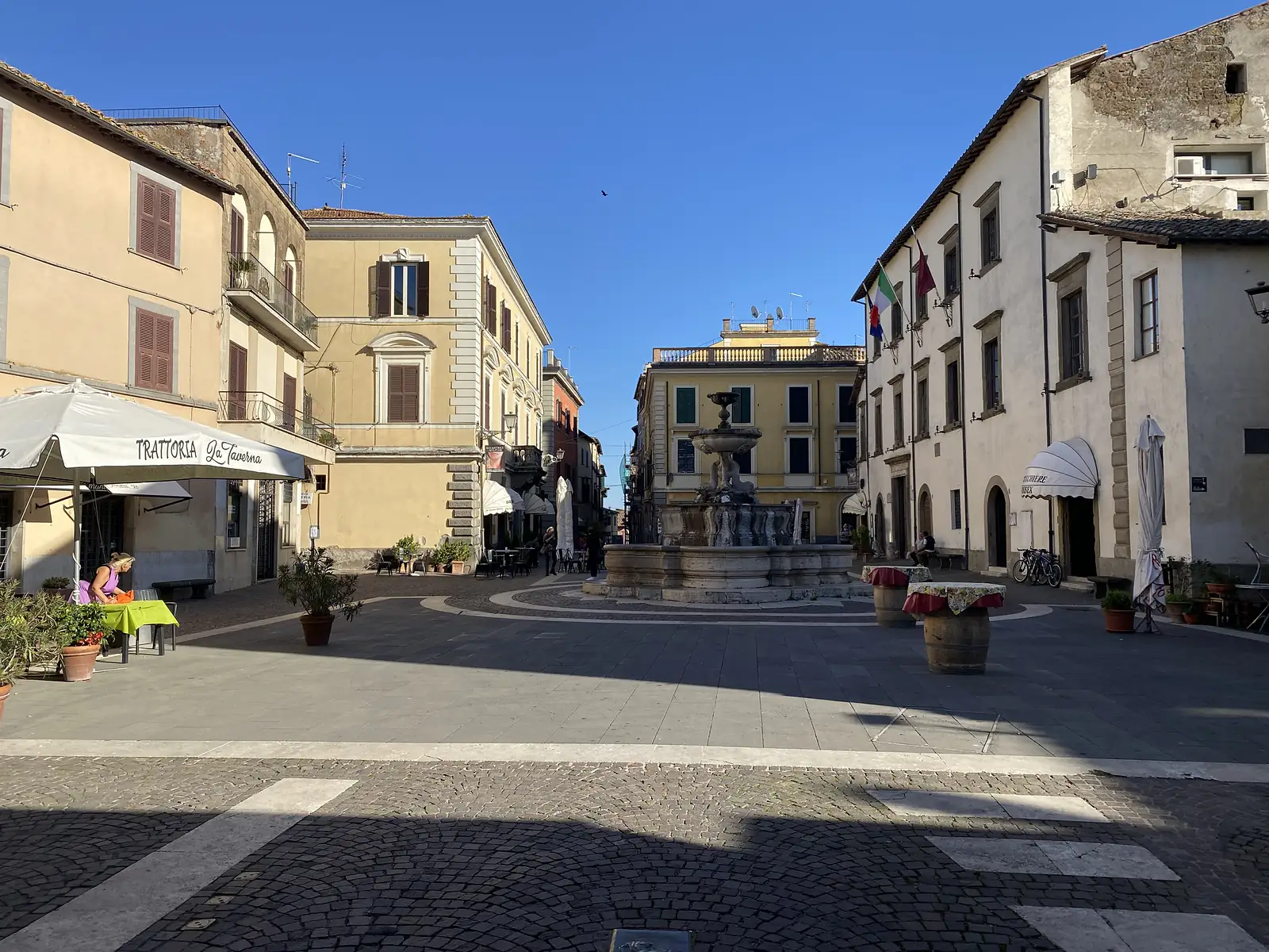 A quiet cobbled town square with a tiered stone fountain at its centre, ochre buildings around it, and outdoor tables outside "Trattoria La Taverna" beneath a white awning.