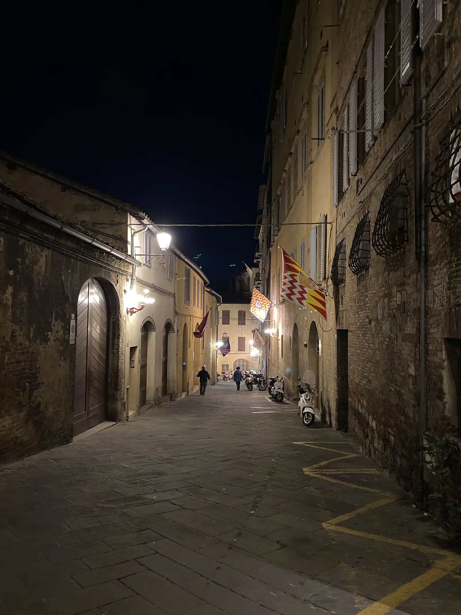 A narrow street at night, its old buildings hung with colourful triangular flags and lit by streetlamps, scooters parked along the wall.