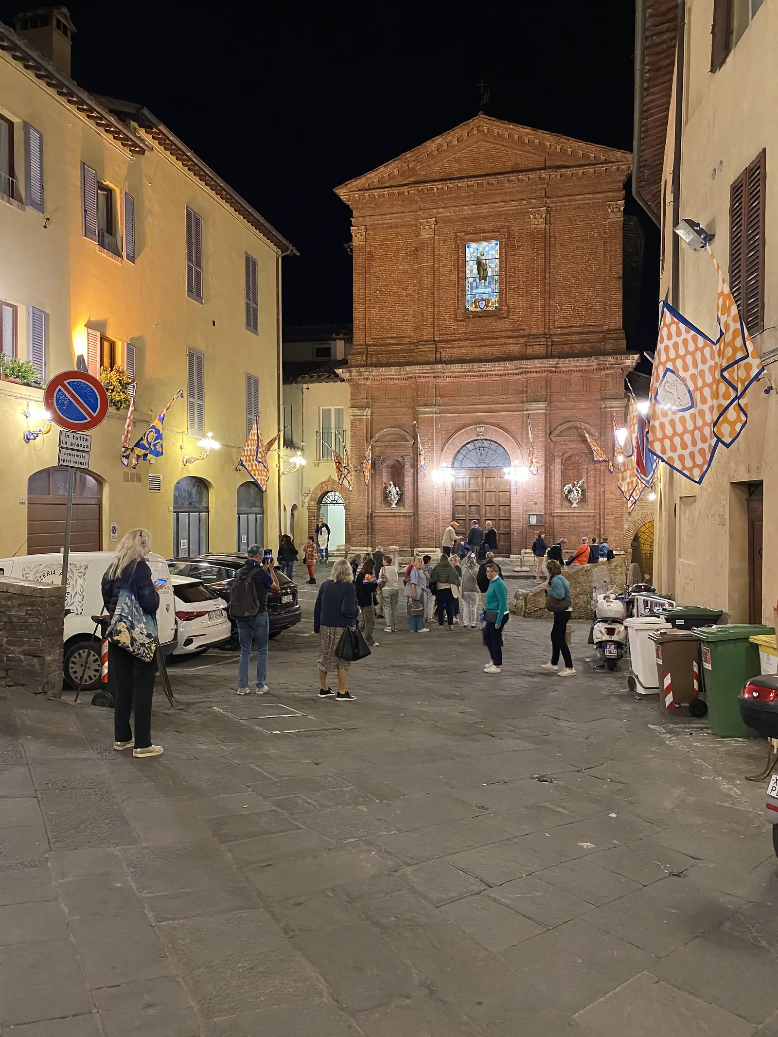 A crowd gathered at night in a piazza before a small brick chapel decorated with a mosaic panel and strings of coloured flags.