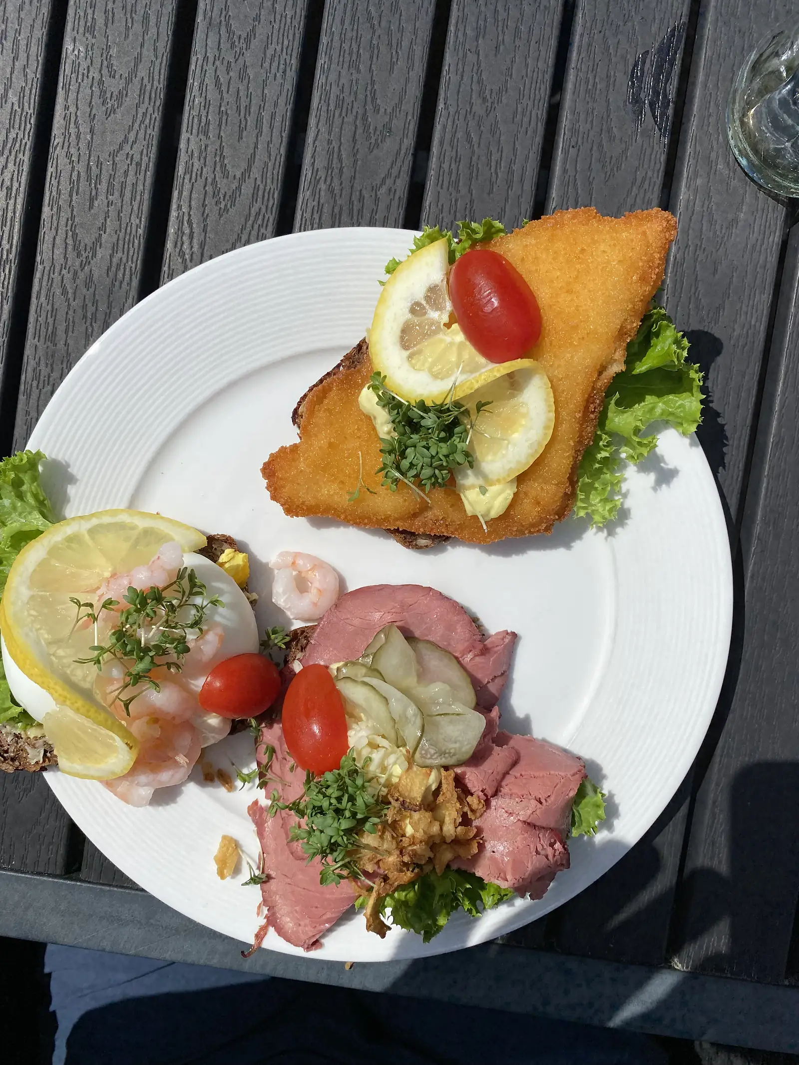 Three open sandwiches on a white plate: breaded fried fish with lemon and cress, prawns with egg, and rare roast beef with pickled cucumber and crispy onions.