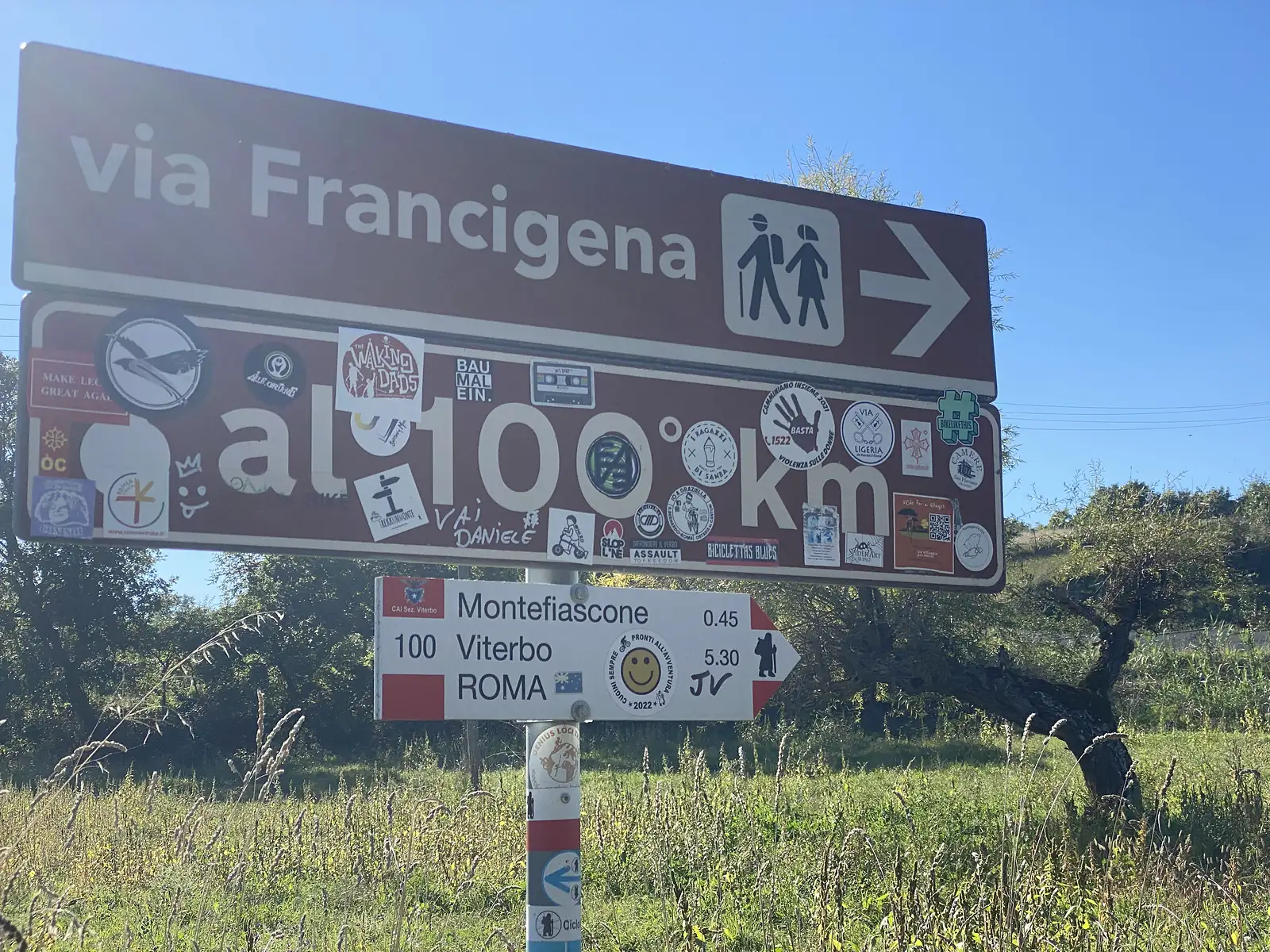 A sticker-covered brown Via Francigena waymark reads "al 100 km", above a white sign pointing to Montefiascone, Viterbo and Roma.