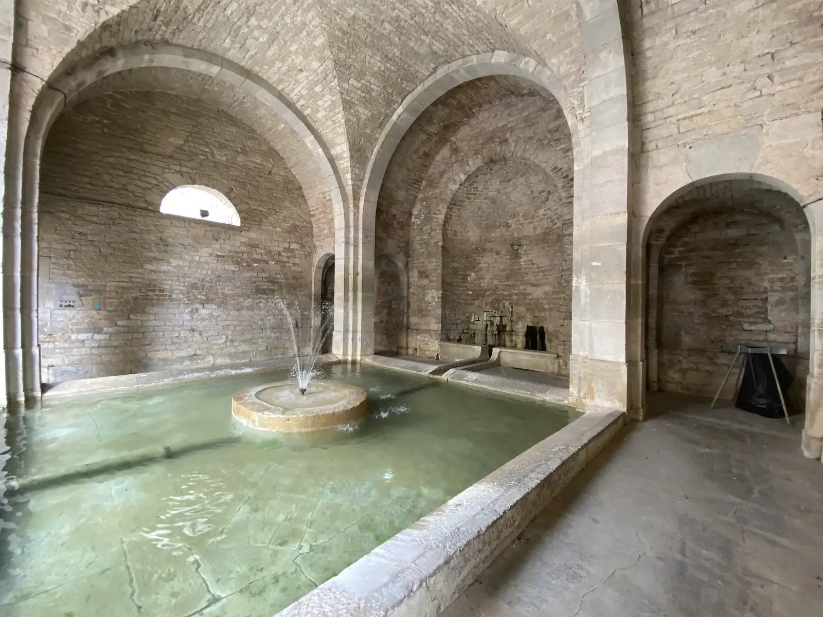 A vaulted stone lavoir with a central fountain jet stands beneath arched brick ceilings, its pool tinged green.