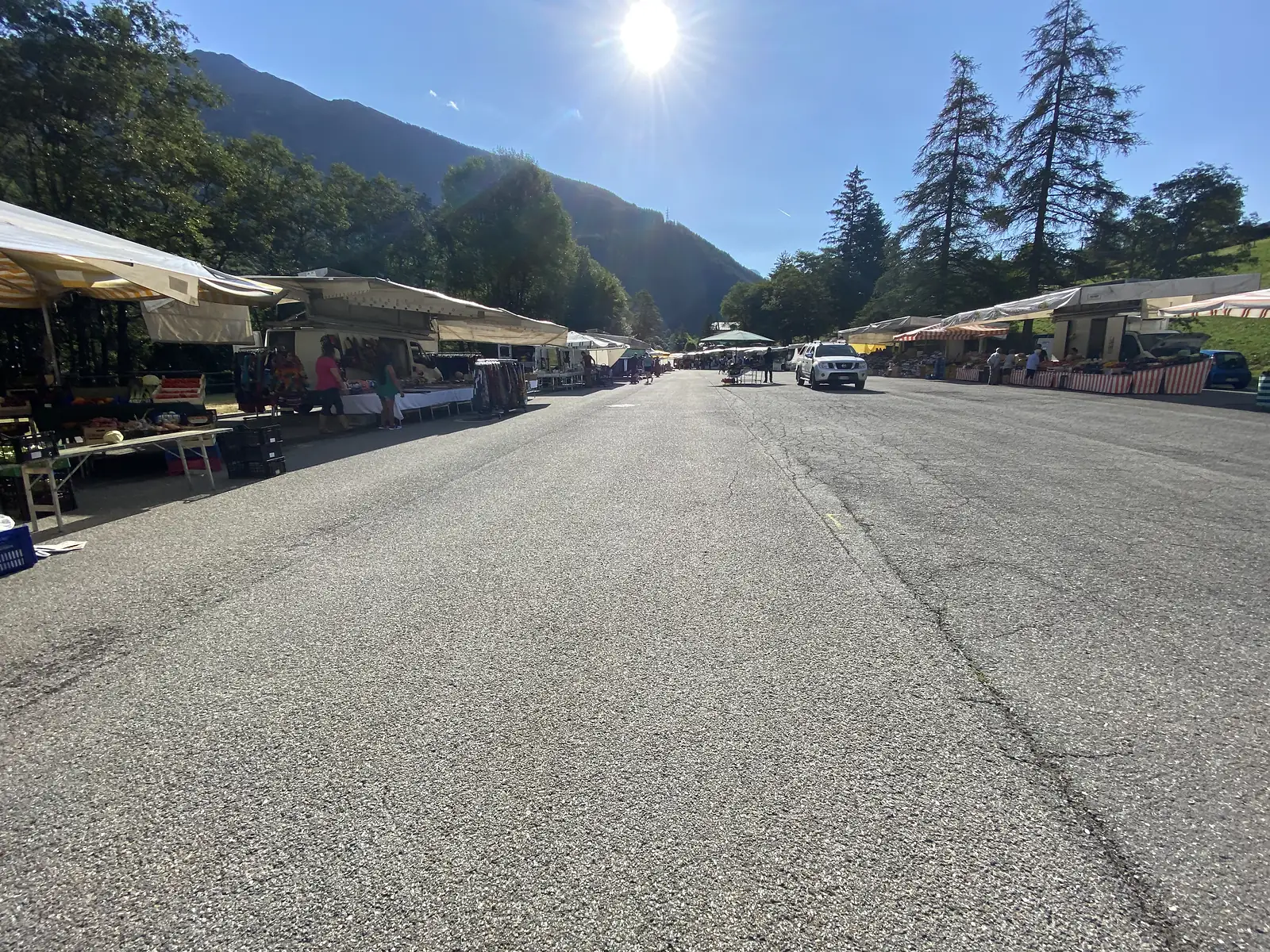 Market stalls with striped awnings line a mountain village car park, shoppers browsing produce and clothing rails with the morning sun low over the surrounding peaks.