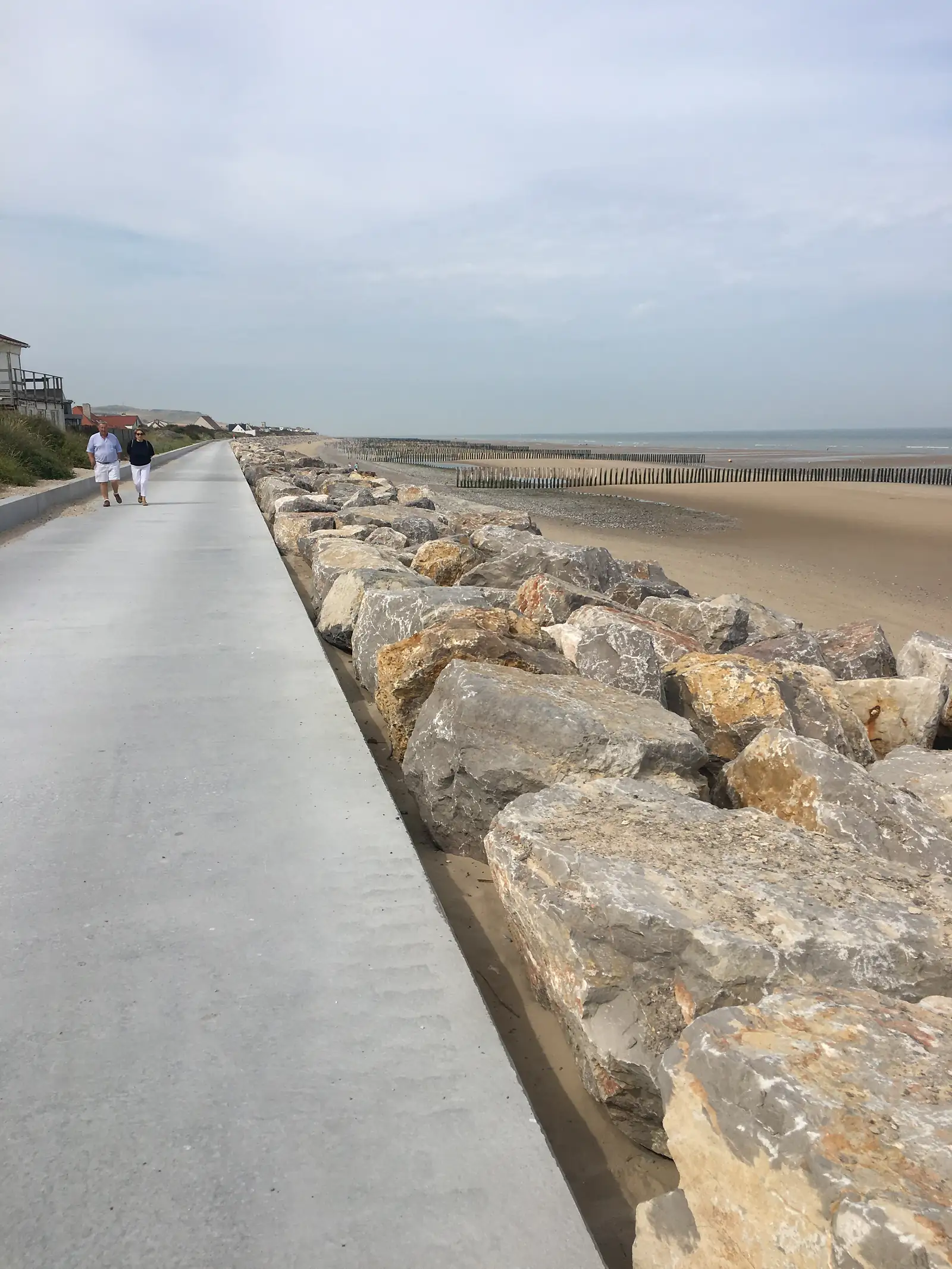 Two walkers in white tops following a concrete promenade beside a line of large rock-armour boulders protecting the shoreline, with sand and sea beyond.