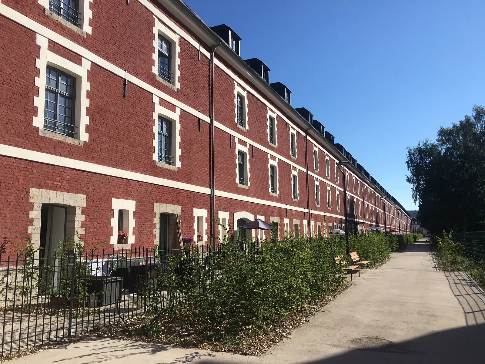 A long terrace of restored red-brick barracks buildings with white dormer windows, overlooking a hedged garden path in Arras.