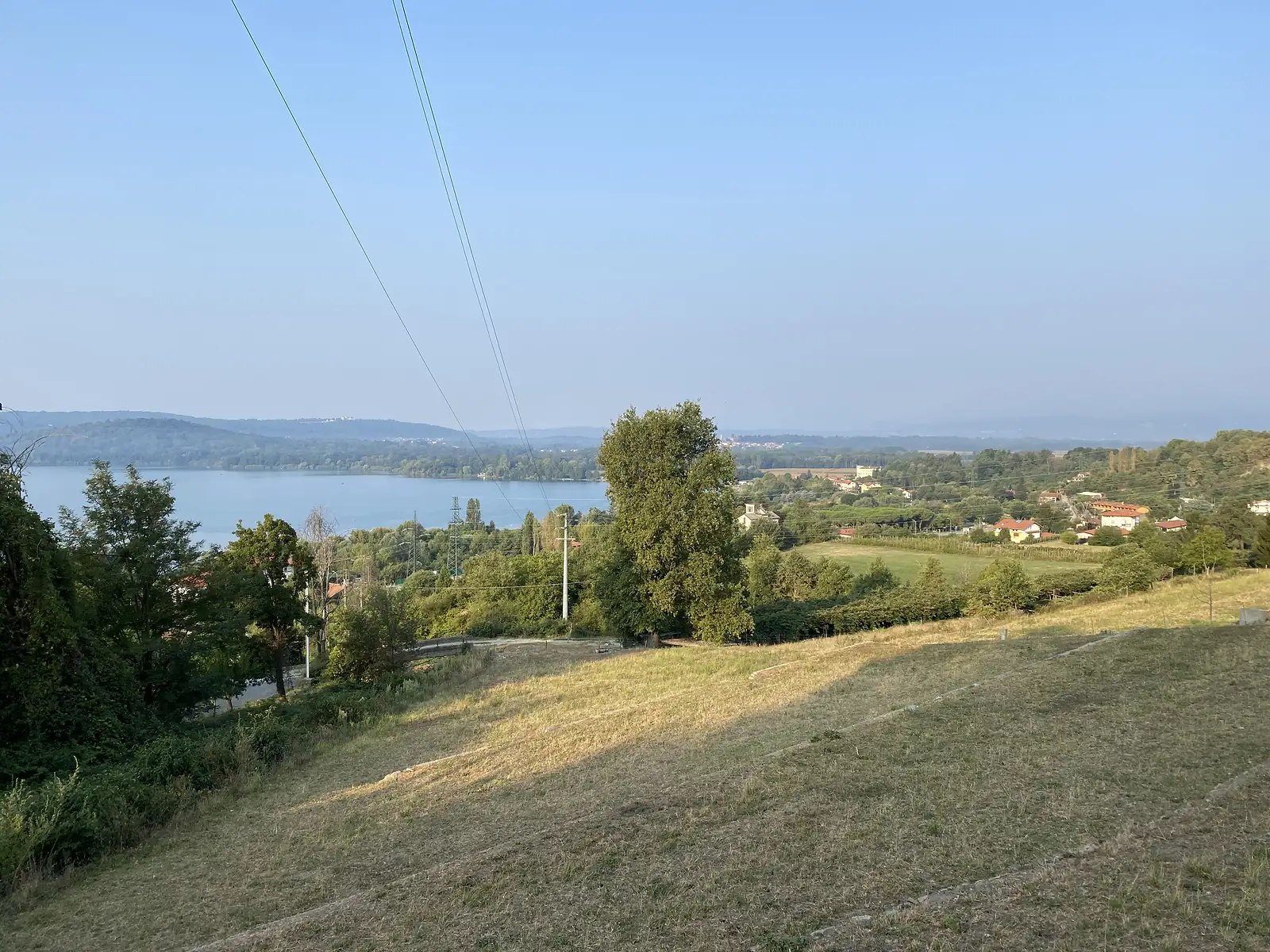 A wide lake seen from a hillside, with a lakeside village and power lines crossing the valley below.