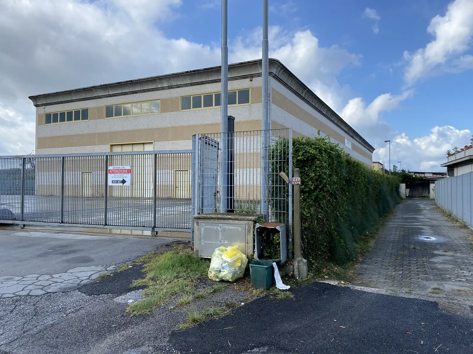 A fenced industrial warehouse with a red 'Attenzione!' sign reading 'General S.R.L., Via delle Ciocche 1157, 300 MT', recycling bins and bin bags left beside the gate.