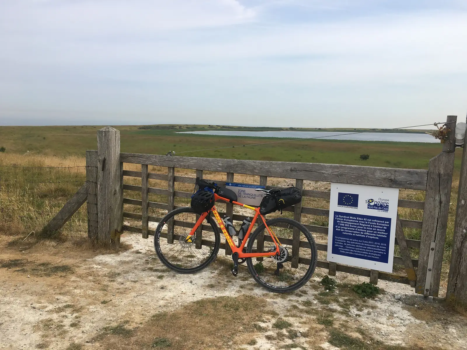 A laden orange gravel bike leaning against a wooden farm gate marked 'Zone de tranquillité de la faune', with a shallow lake and open grassland beyond.