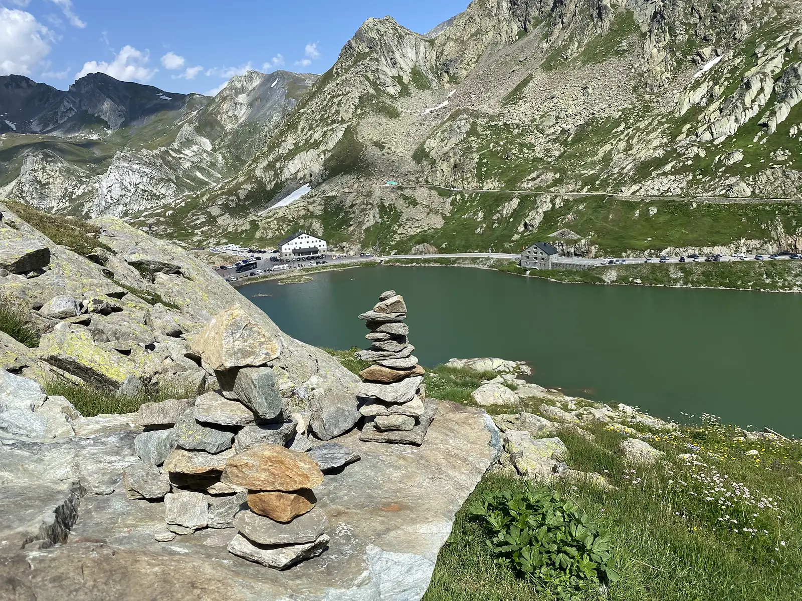 Two small cairns of balanced stones on a rock ledge overlooking the green pass lake, the hospice and a line of parked cars on its far bank.