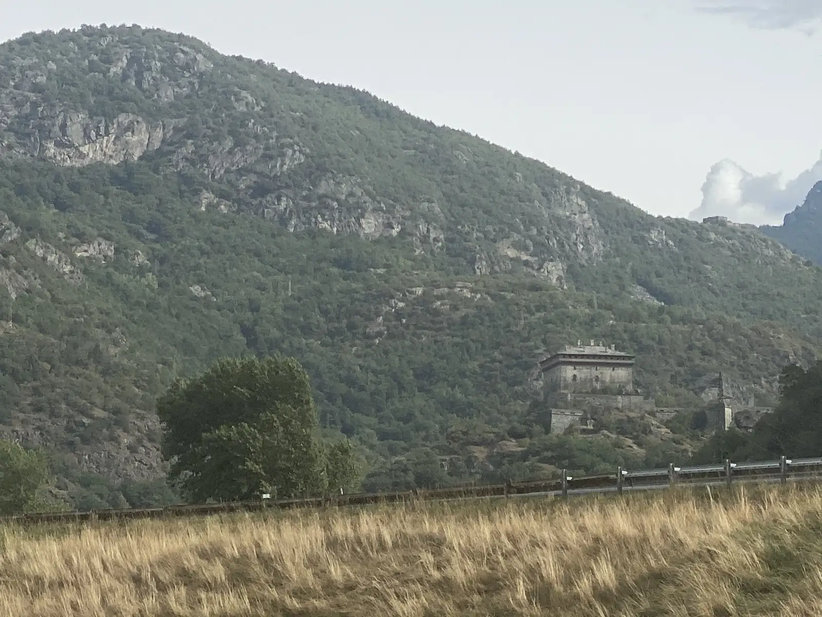 A fortified stone building perched on a rocky spur partway up a steep wooded mountainside, seen across a grassy field bordered by a guardrail.