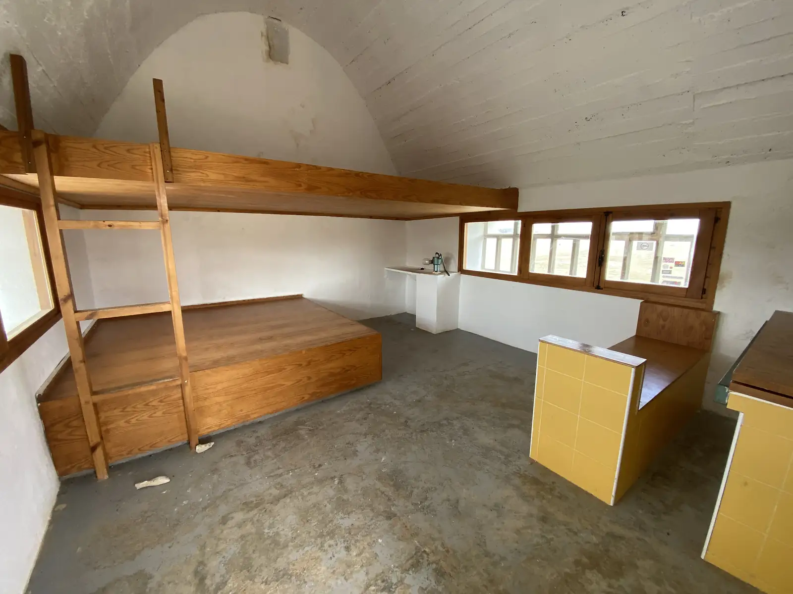 The bare whitewashed interior of a mountain refuge, with wooden sleeping platforms, a bunk ladder and a yellow-tiled counter under a barrel-vaulted roof.