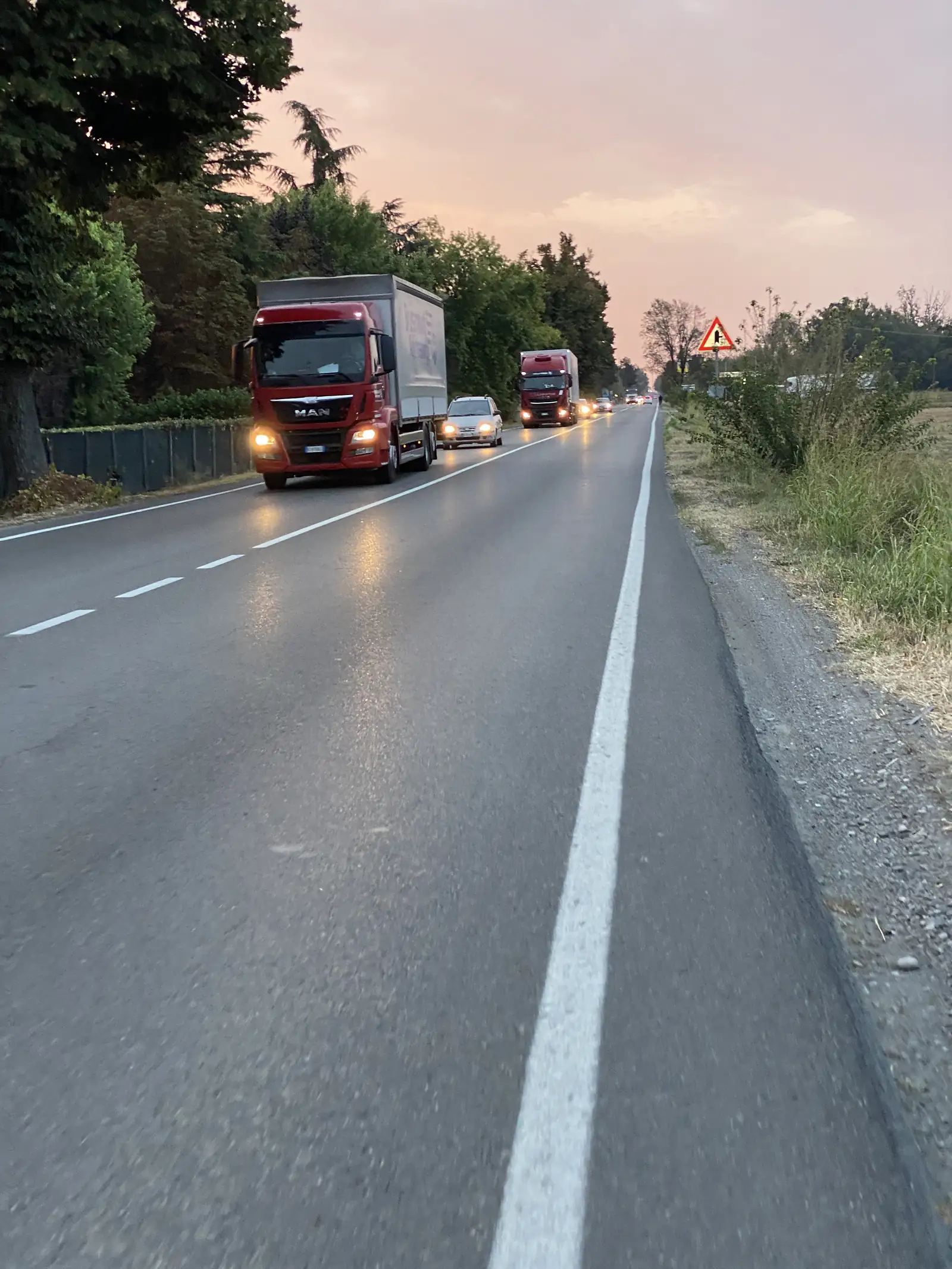 A line of red MAN lorries with headlights on, queued on a rural road at dusk beside a triangular warning sign.
