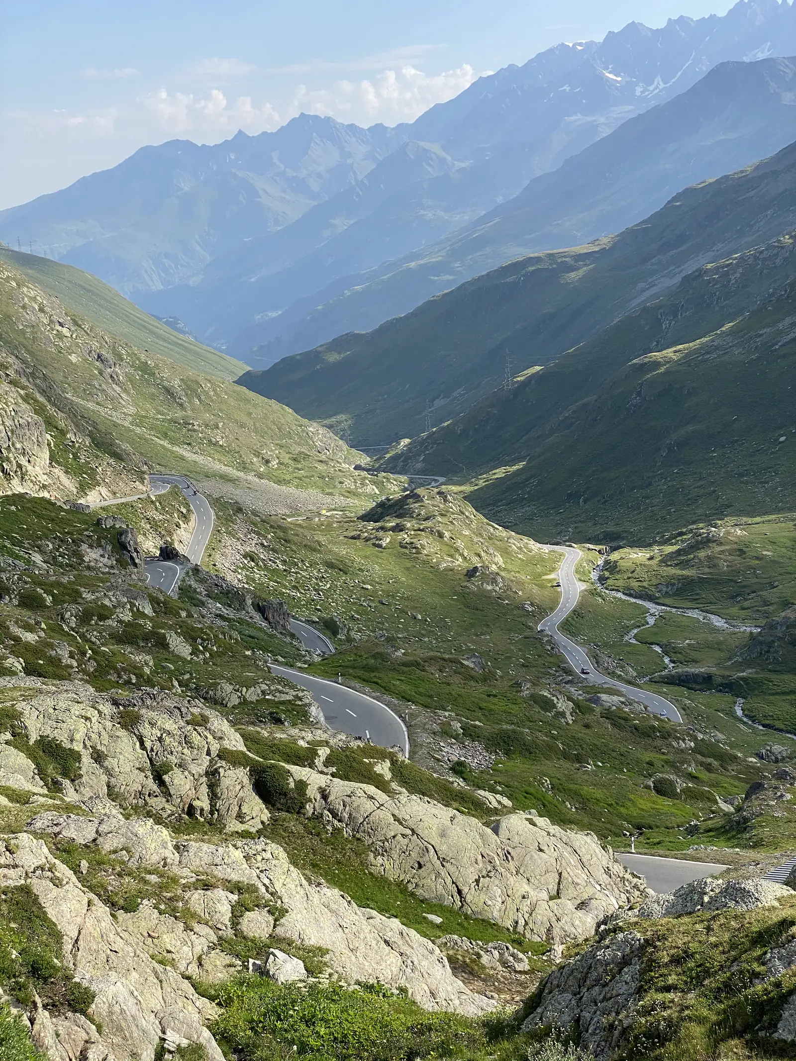 A mountain road twists down through a valley in tight switchbacks, with jagged snow-streaked peaks in the distance.