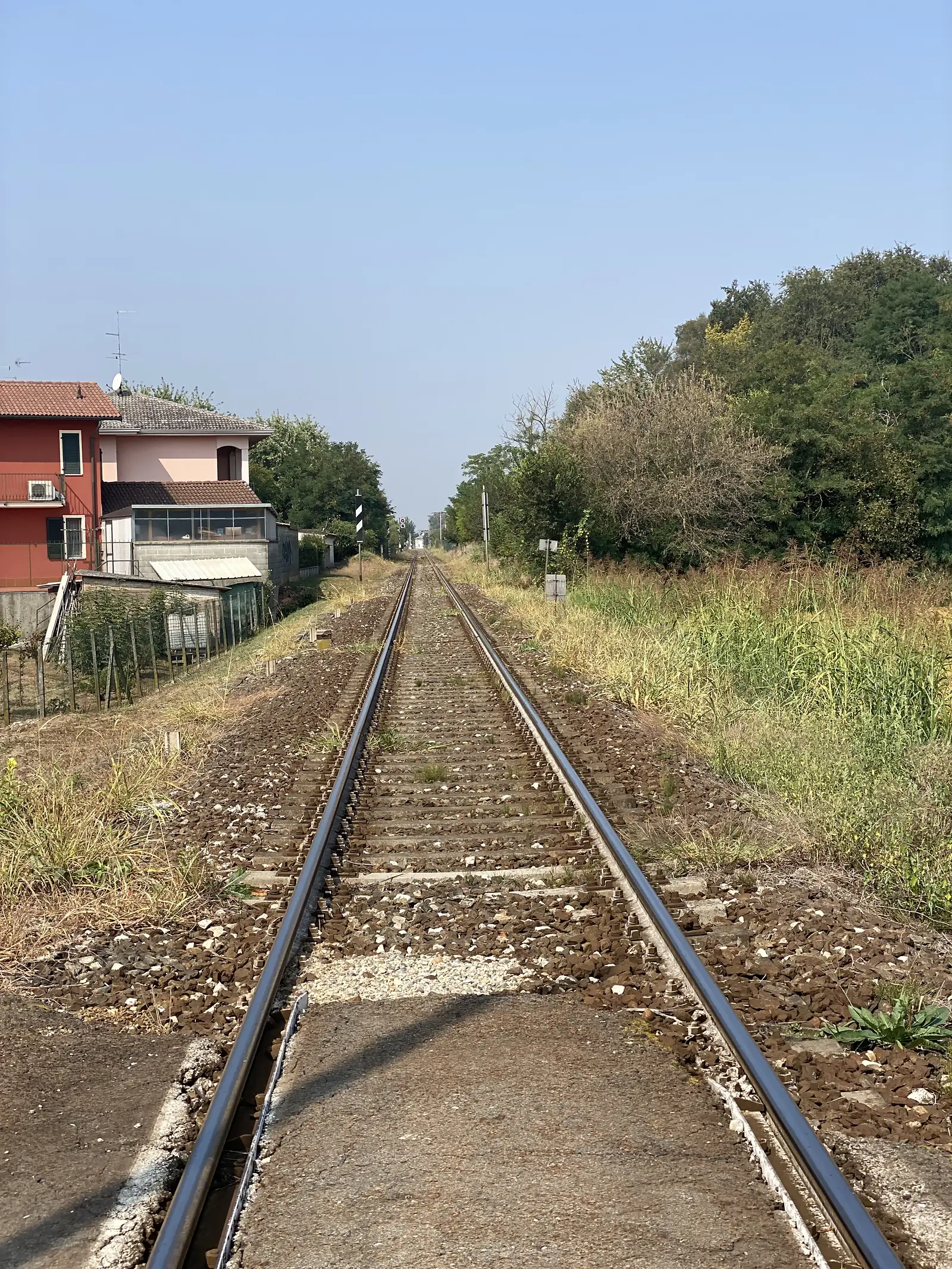 A single railway track runs straight past a level crossing towards a red-roofed house, flanked by trees and rough grass.